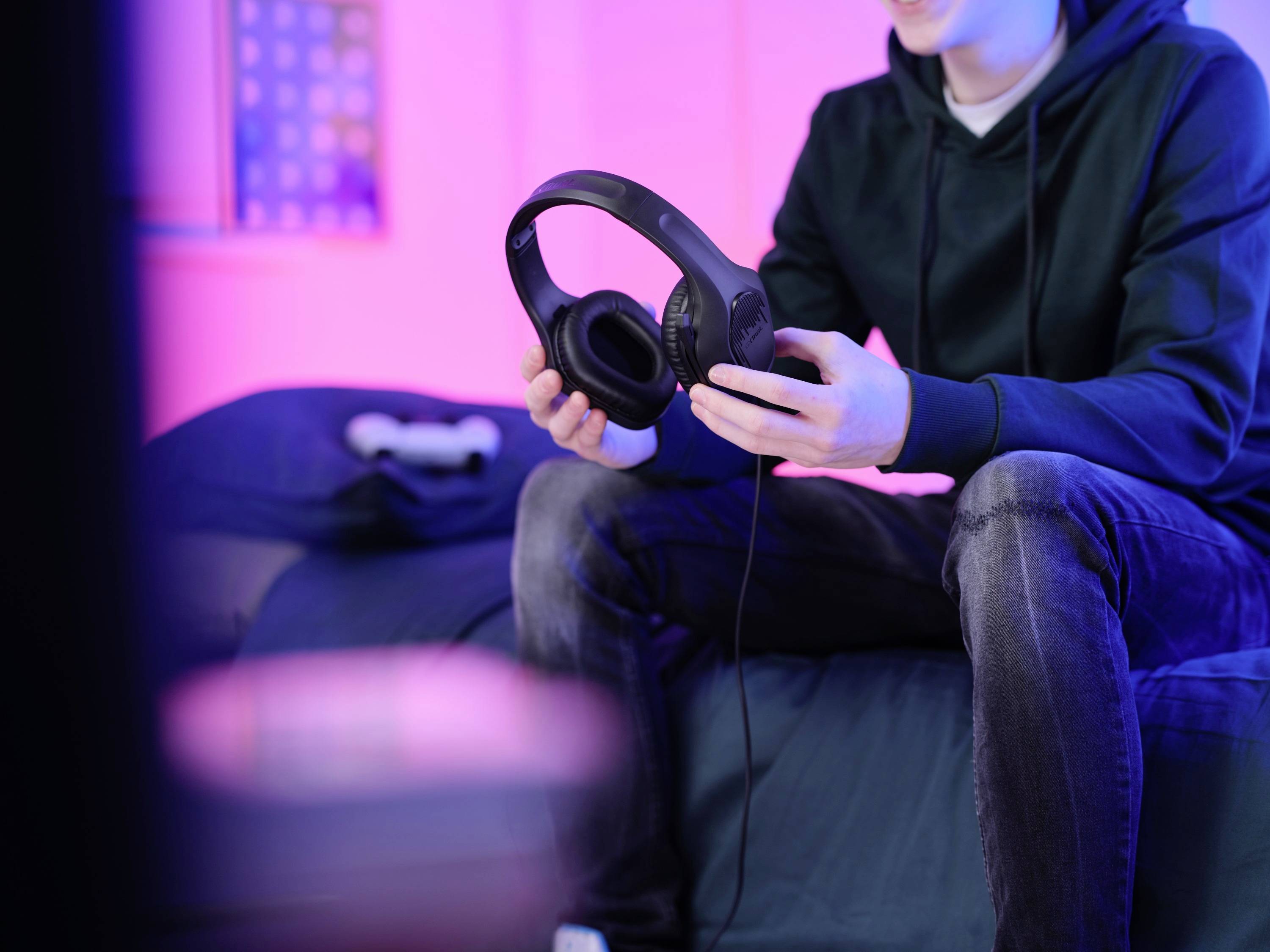 Eine Person sitzt auf einem Bett in einem Zimmer mit pinker Beleuchtung und hält einen schwarzen Kopfhörer. Im Hintergrund ist ein Controller sichtbar.