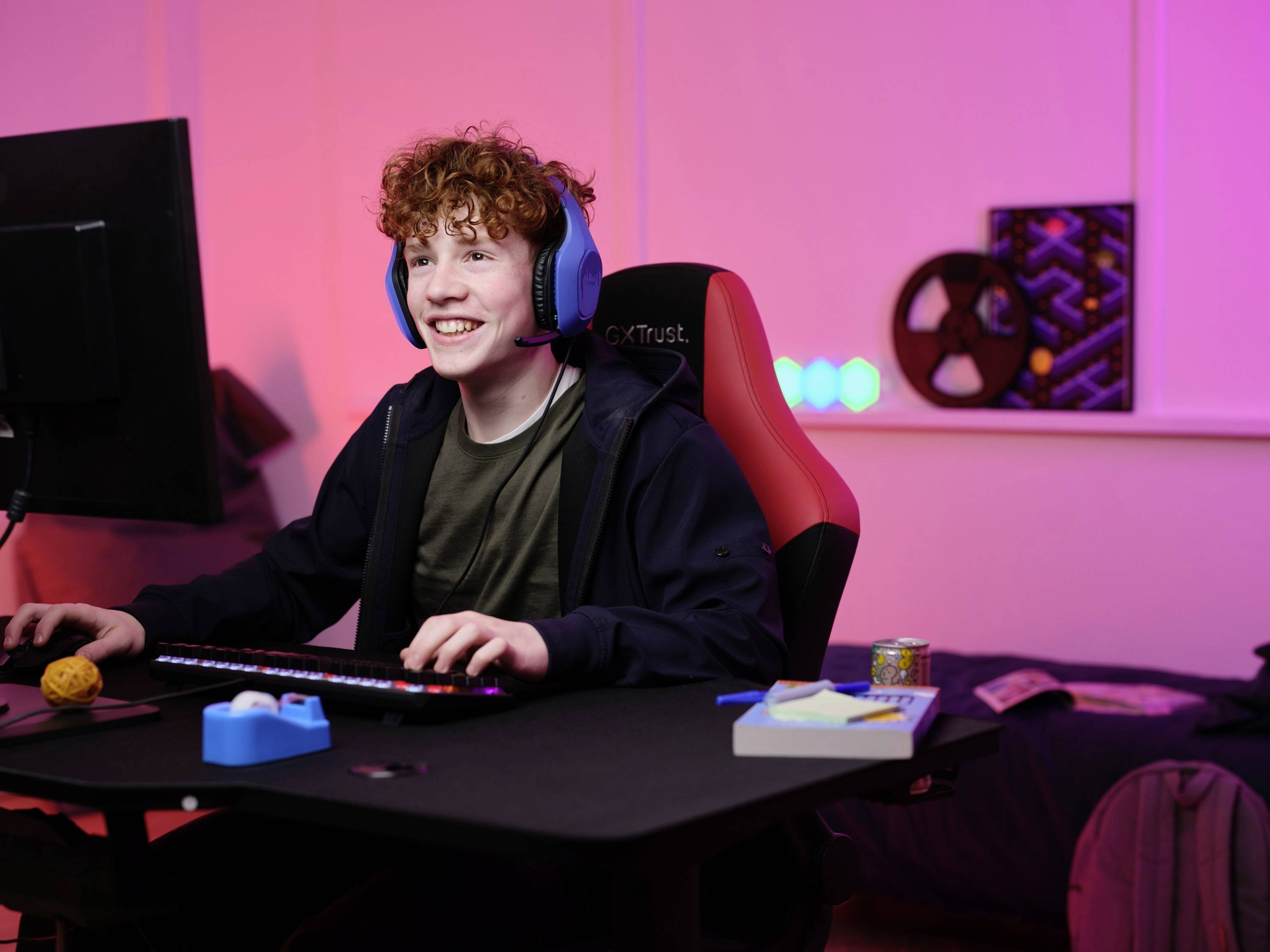 Ein Jugendlicher sitzt lächelnd mit Kopfhörern vor einem PC in einem modern eingerichteten Zimmer mit einer bunten Beleuchtung.