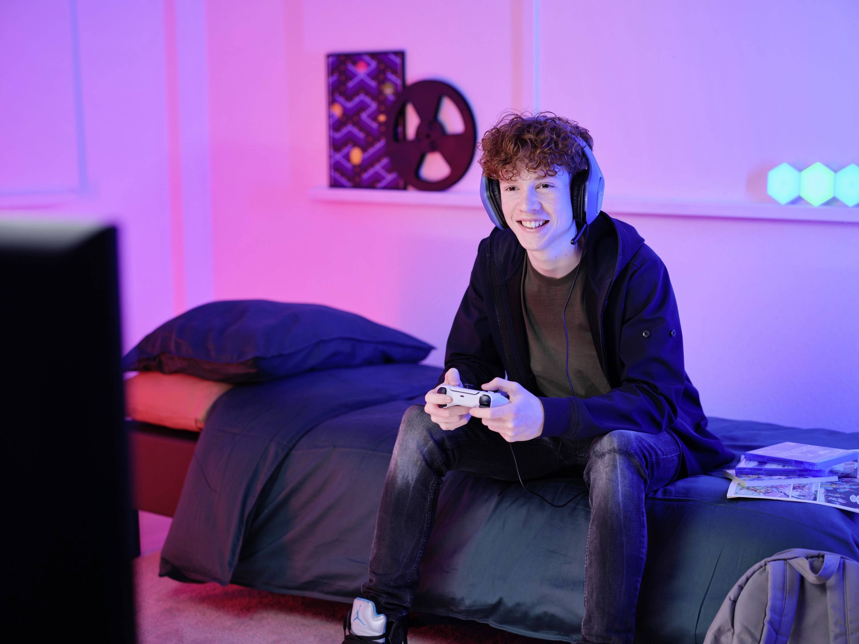 Junger Mann sitzt auf einem Bett mit Kopfhörern und spielt mit einem Gamecontroller. Im Hintergrund bunte Lichter und ein dekoratives Element.