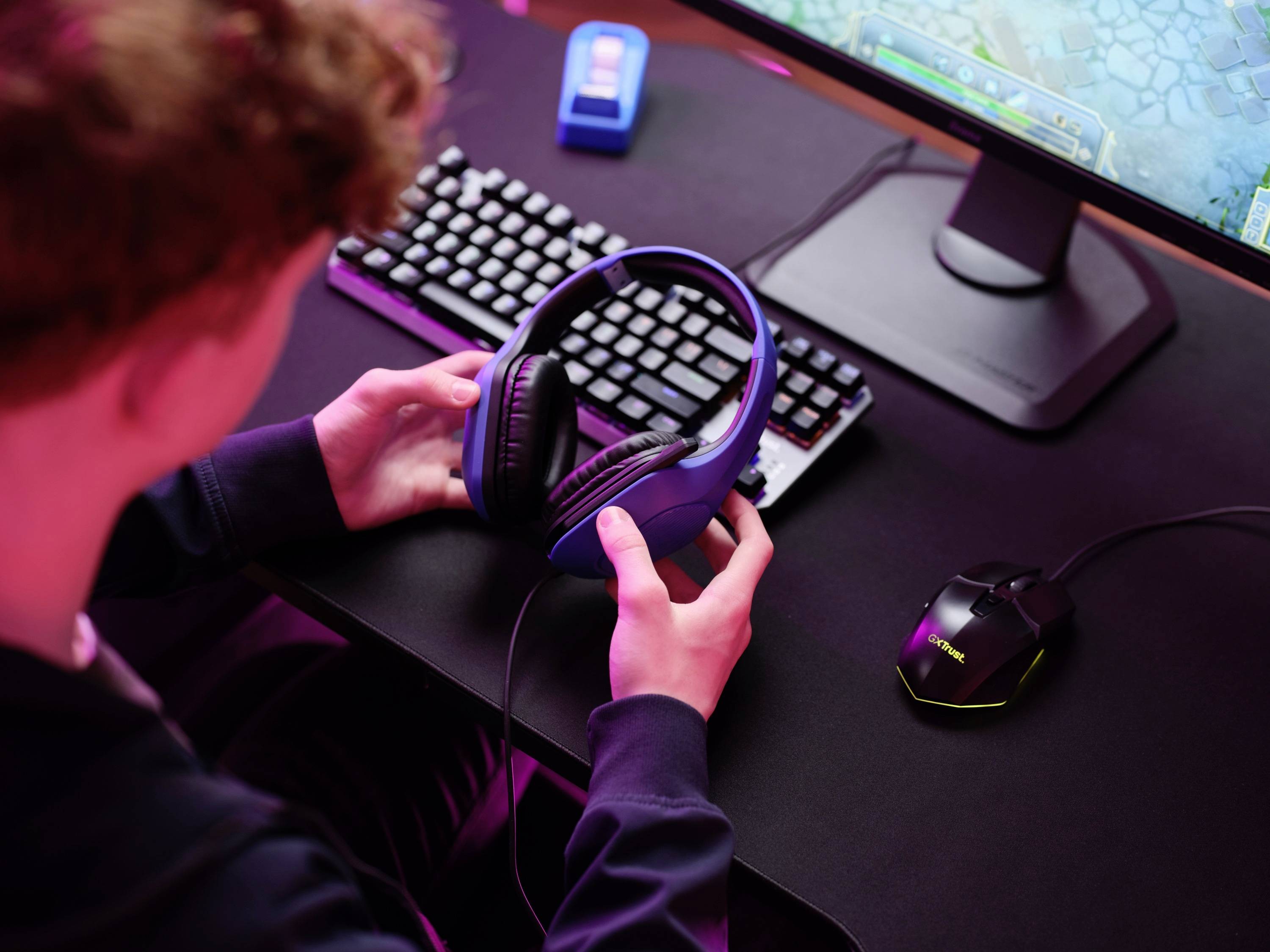 Eine Person sitzt an einem Schreibtisch mit Computer, hält Kopfhörer. Der Bildschirm zeigt ein Videospiel.