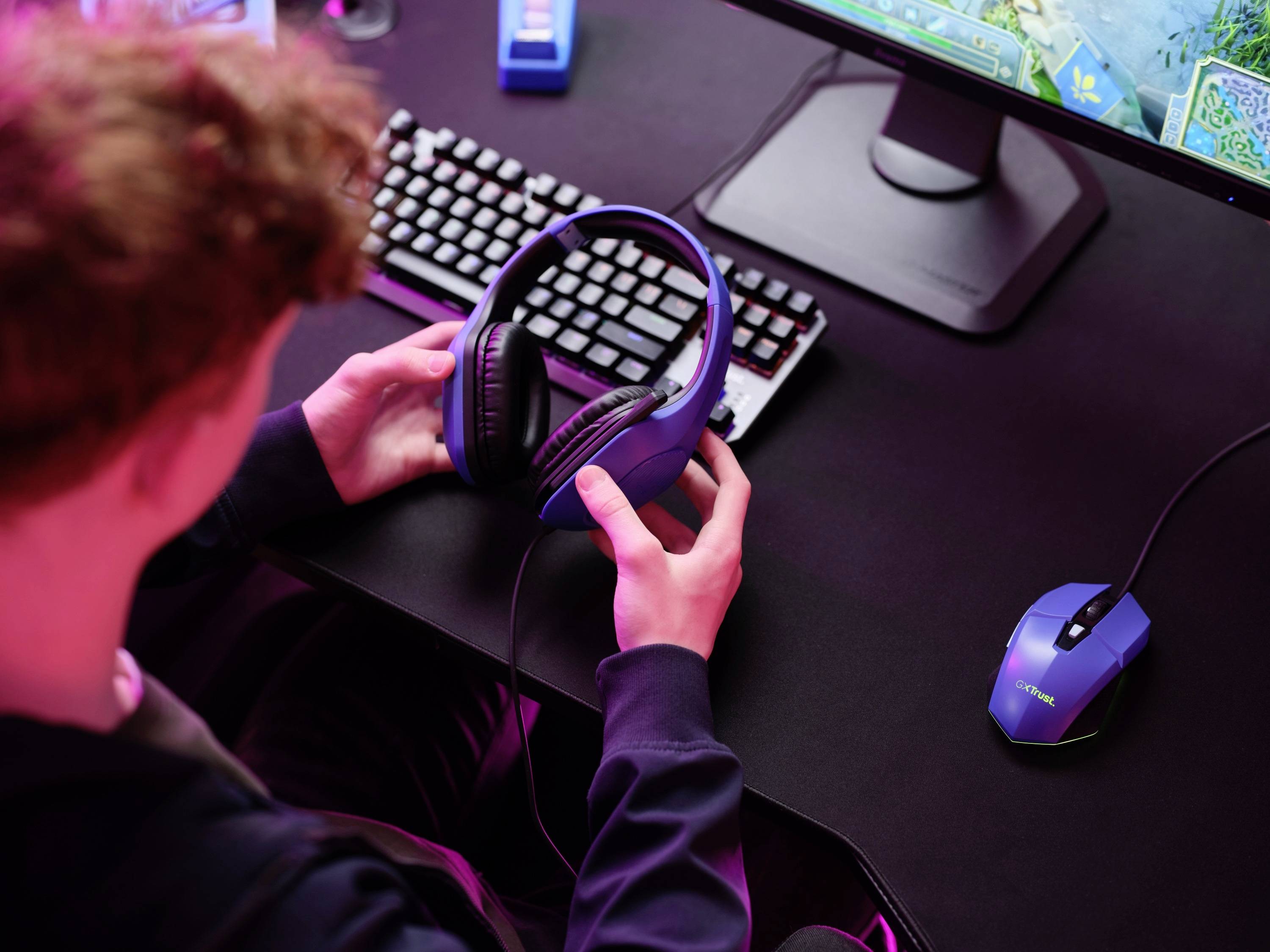 Eine Person hält Kopfhörer vor einem Computerbildschirm und Tastatur. Umgebung ist von rosa Licht beleuchtet.