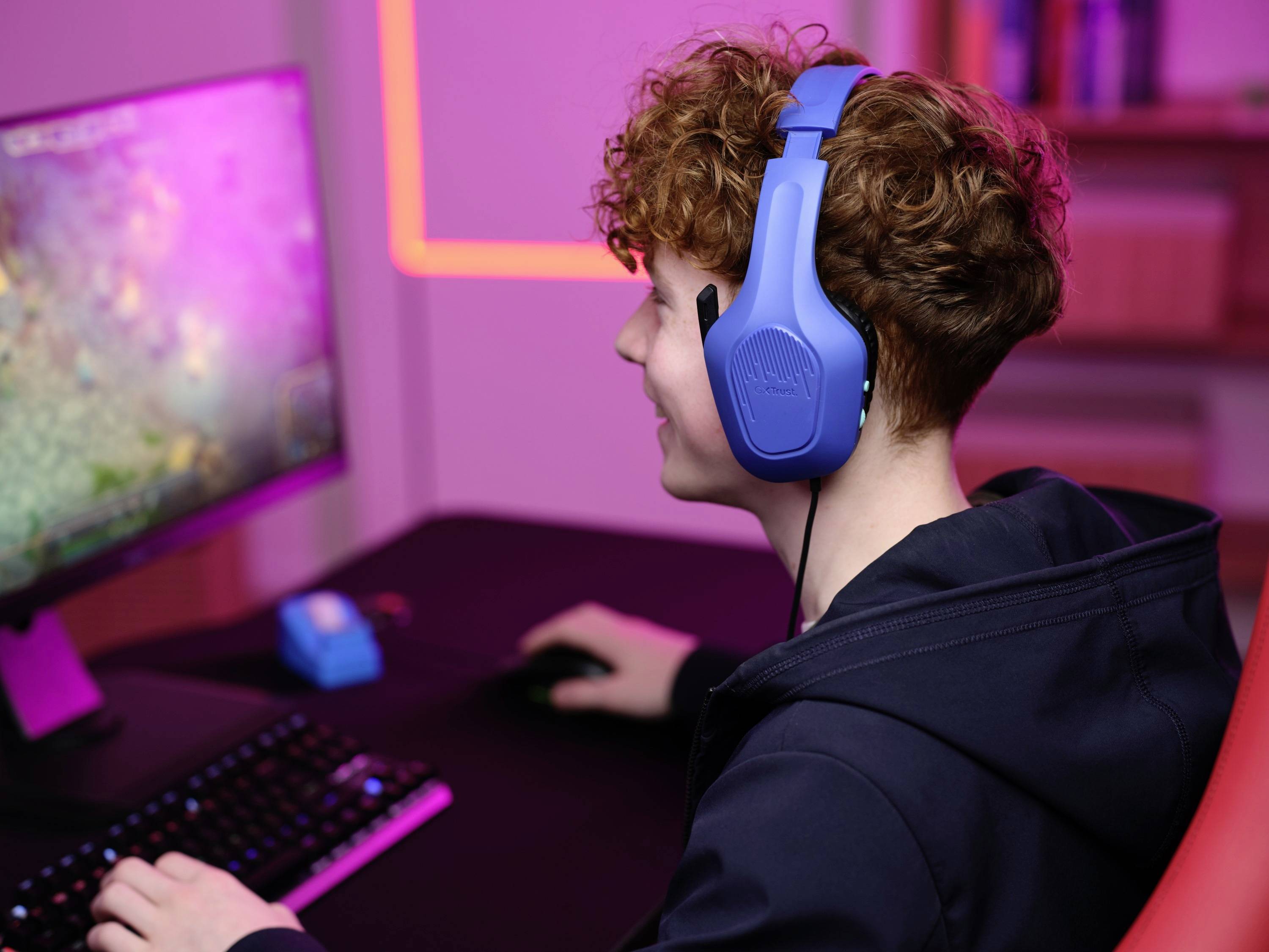 Person mit blauen Kopfhörern sitzt an einem Schreibtisch vor einem Monitor und tippt auf einer Tastatur, umgeben von rosa Licht.