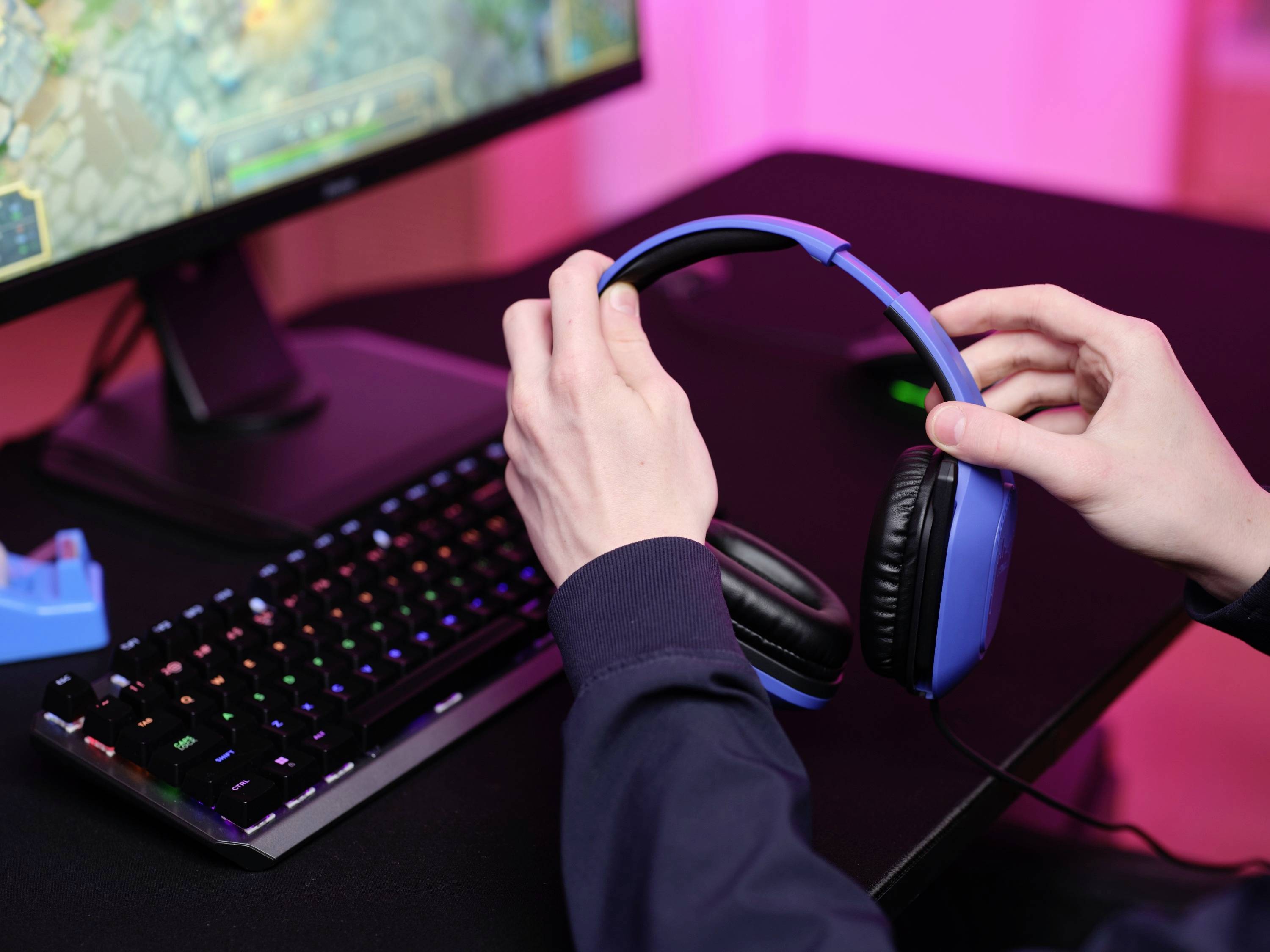 Eine Person legt ein blaues Headset auf einen Schreibtisch neben eine beleuchtete Tastatur und einen Monitor mit einem Spiel.