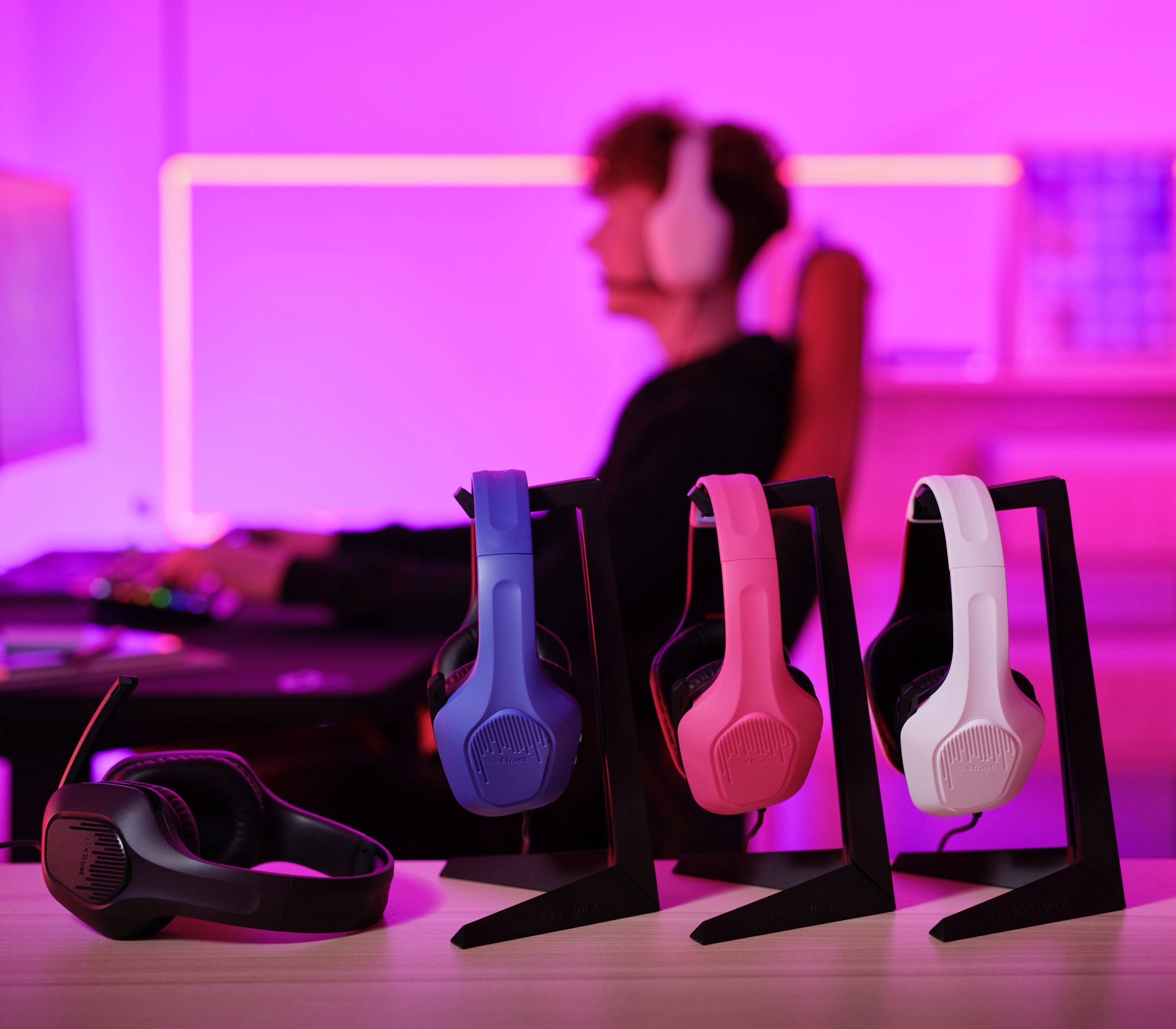 Eine Person trägt ein Headset und sitzt an einem Schreibtisch vor einem Bildschirm. Im Vordergrund stehen drei verschiedenfarbige Headsets.