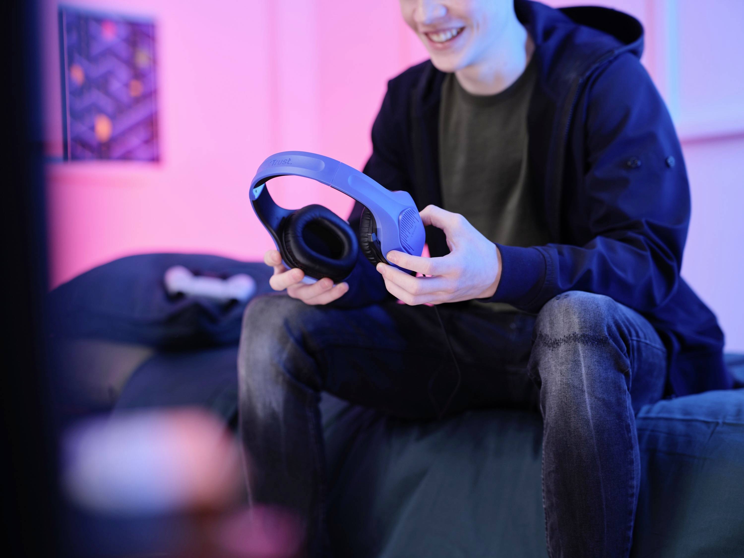 Eine Person sitzt lächelnd auf einem Bett in einem Raum mit pinkem Licht, hält Kopfhörer in den Händen.