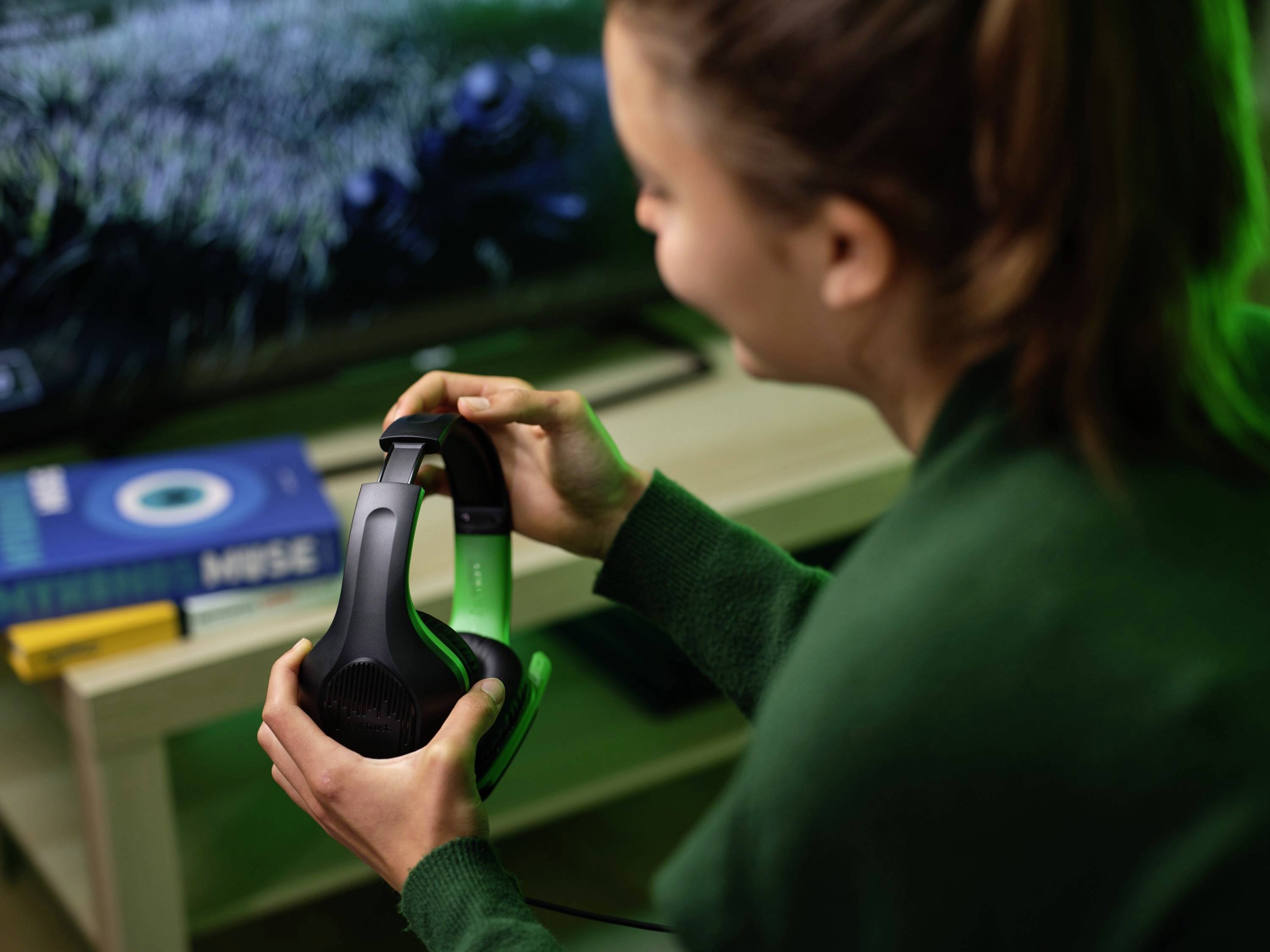 Eine Person hält ein Paar schwarze Kopfhörer in der Hand. Im Hintergrund sind ein Fernseher und Bücher auf einem Tisch sichtbar.