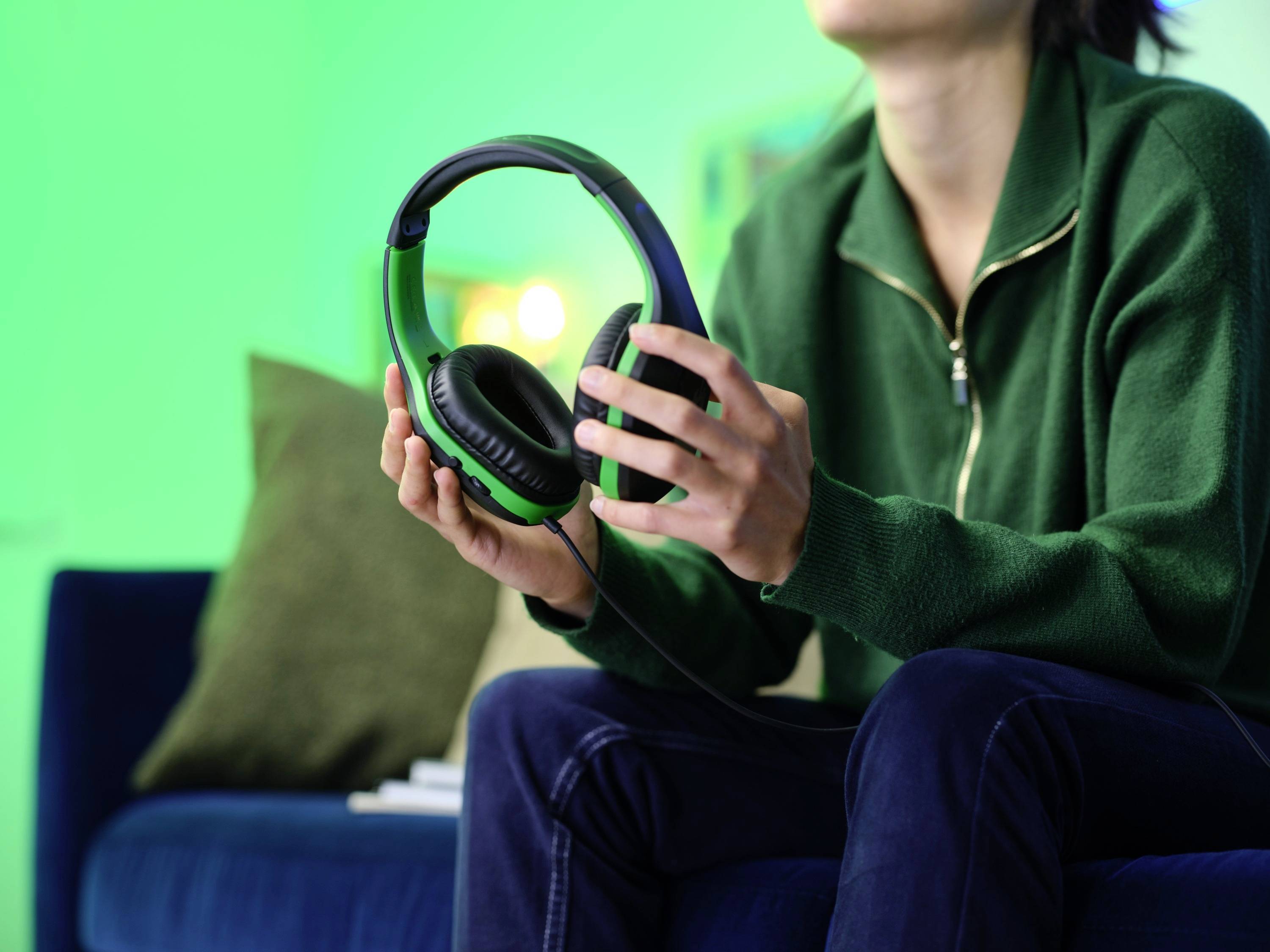 Person auf einem Sofa hält grüne Kopfhörer in der Hand. Hintergrund ist grünlich beleuchtet. Person trägt einen grünen Pullover.