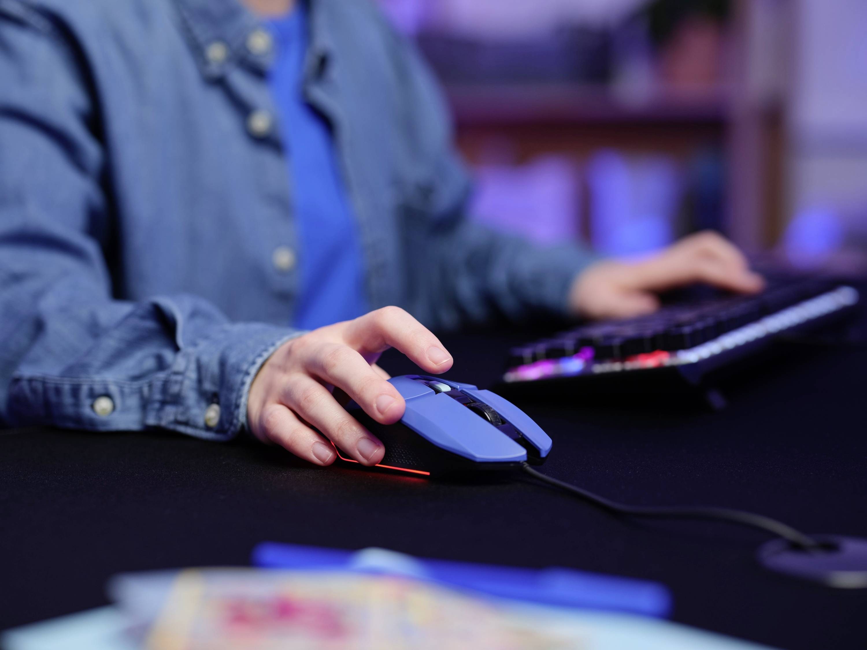 Person in blauer Kleidung benutzt eine Computermaus und Tastatur auf einem Schreibtisch. Büroumgebung im Hintergrund.