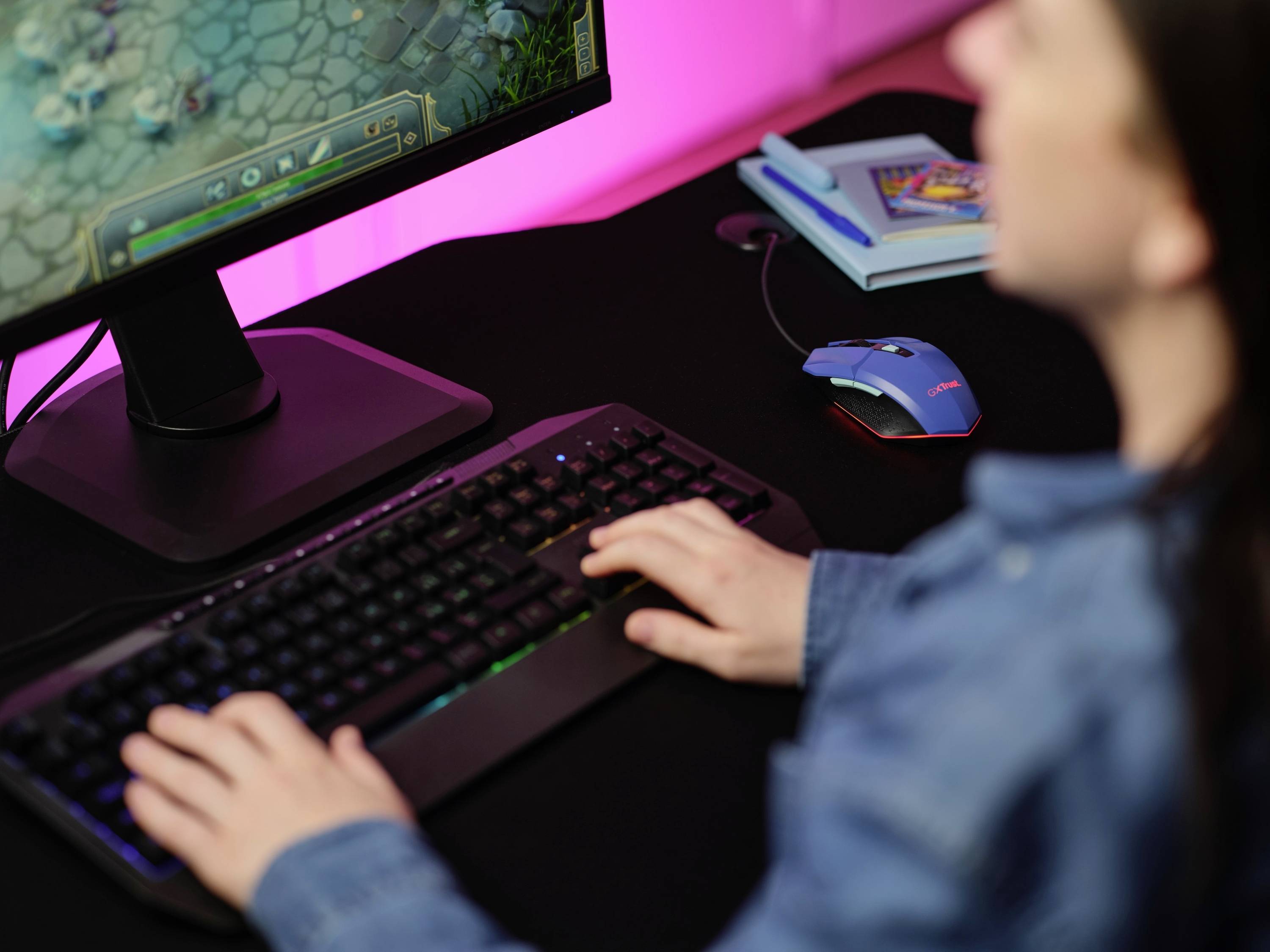 Eine Person spielt ein Computerspiel auf einem Monitor, mit einer Tastatur und Maus auf dem Schreibtisch. Im Hintergrund sind Notizbücher zu sehen.