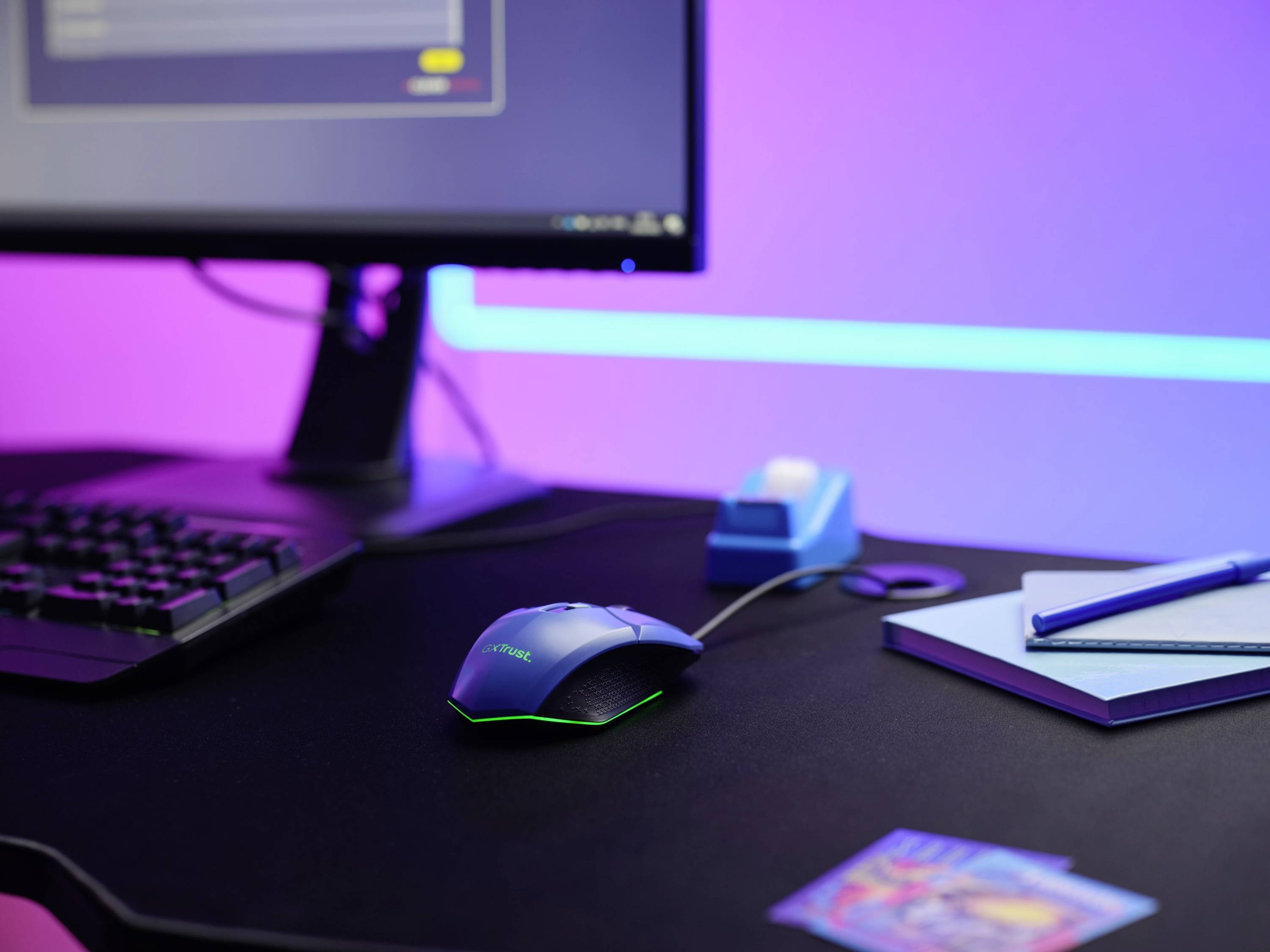 Ein Schreibtisch mit einem Computerbildschirm, Tastatur, Maus und Schreibutensilien unter blau-lila Beleuchtung.