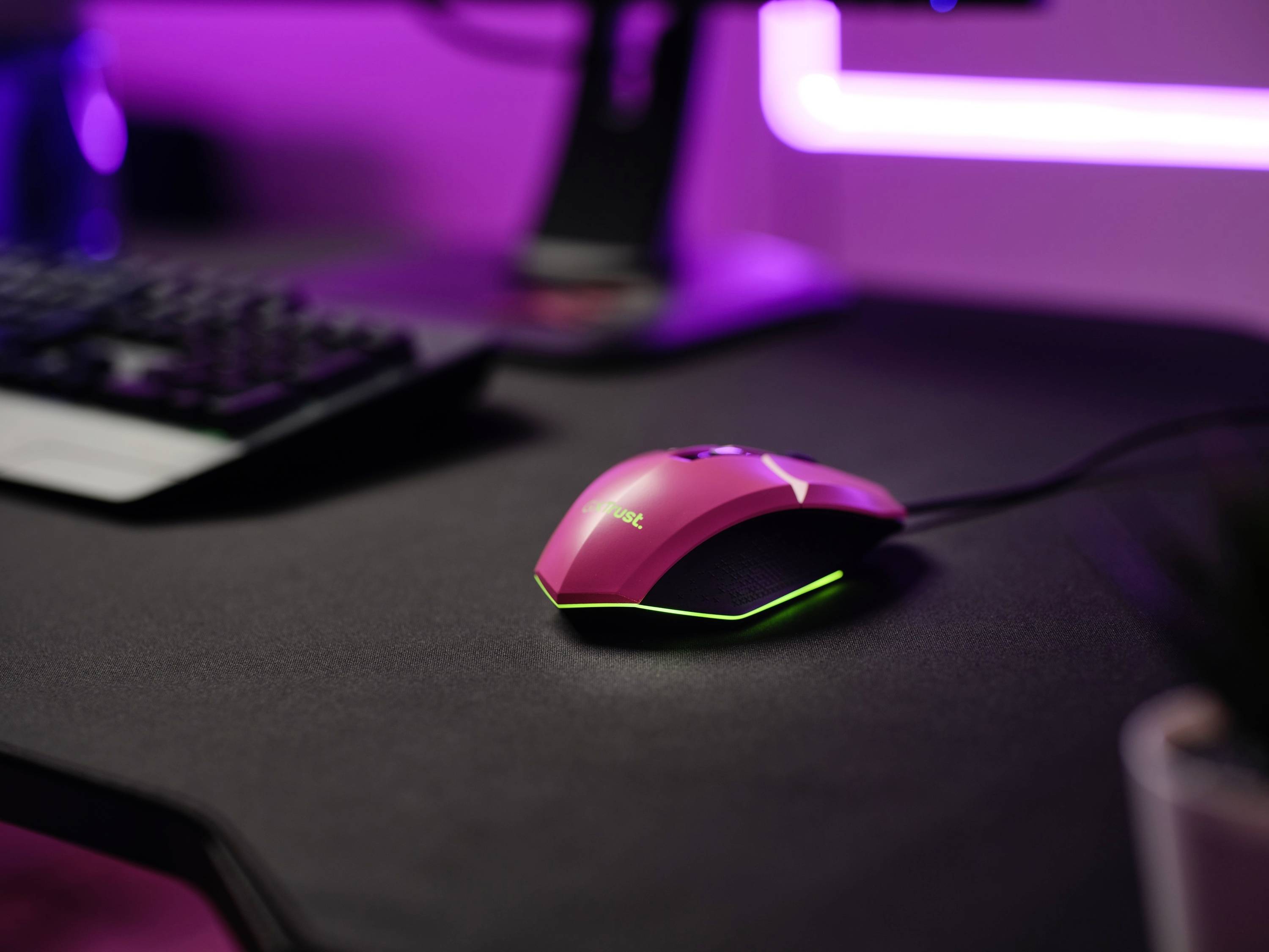 Eine rosa Gaming-Maus mit Beleuchtung liegt auf einem Schreibtisch mit einer Tastatur im Hintergrund, modernes Setup.