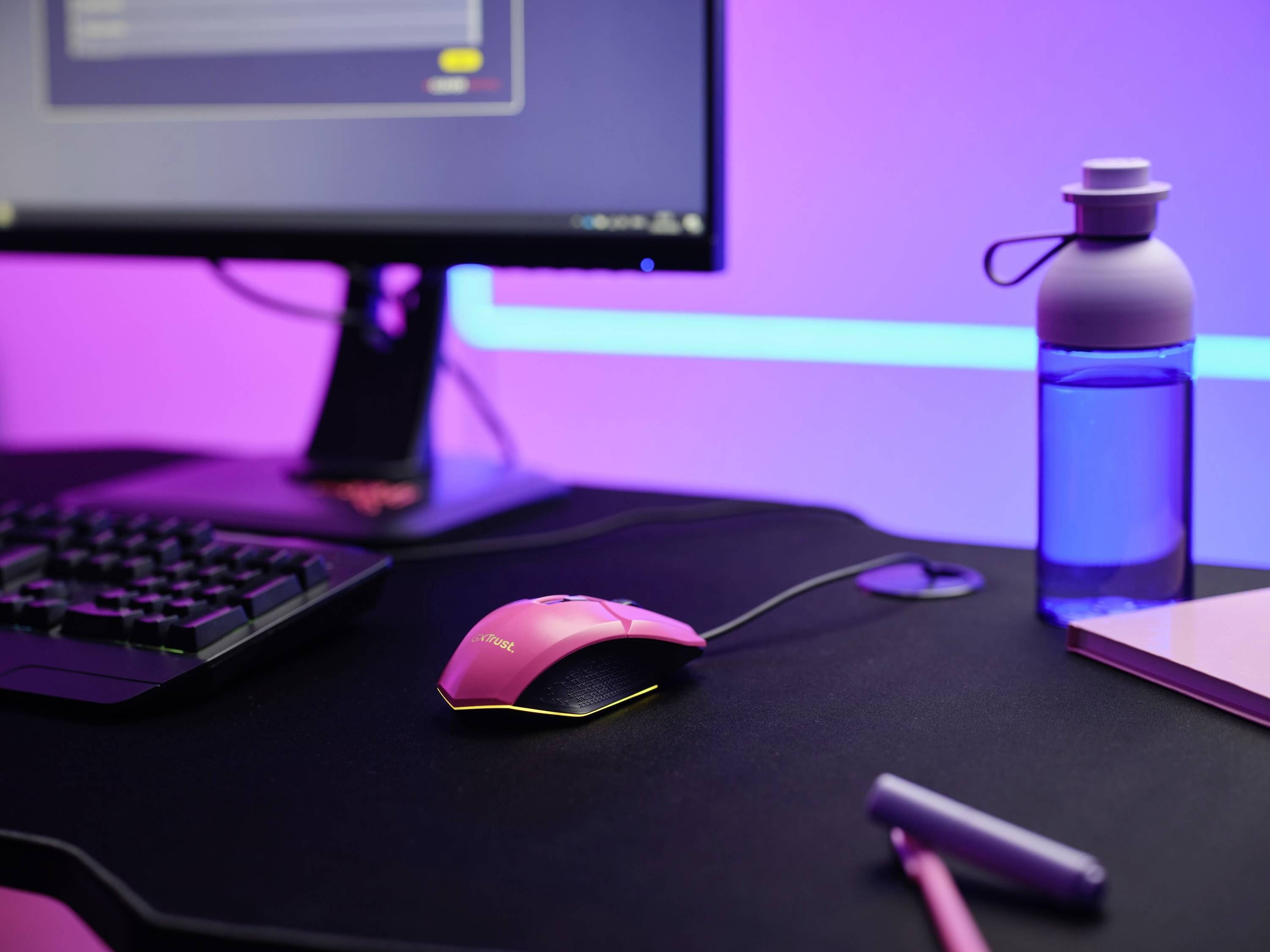 Ein Schreibtisch mit einem rosa Computermaus, einer Tastatur, einem Monitor, einer blauen Wasserflasche und einem rosa Notizbuch.