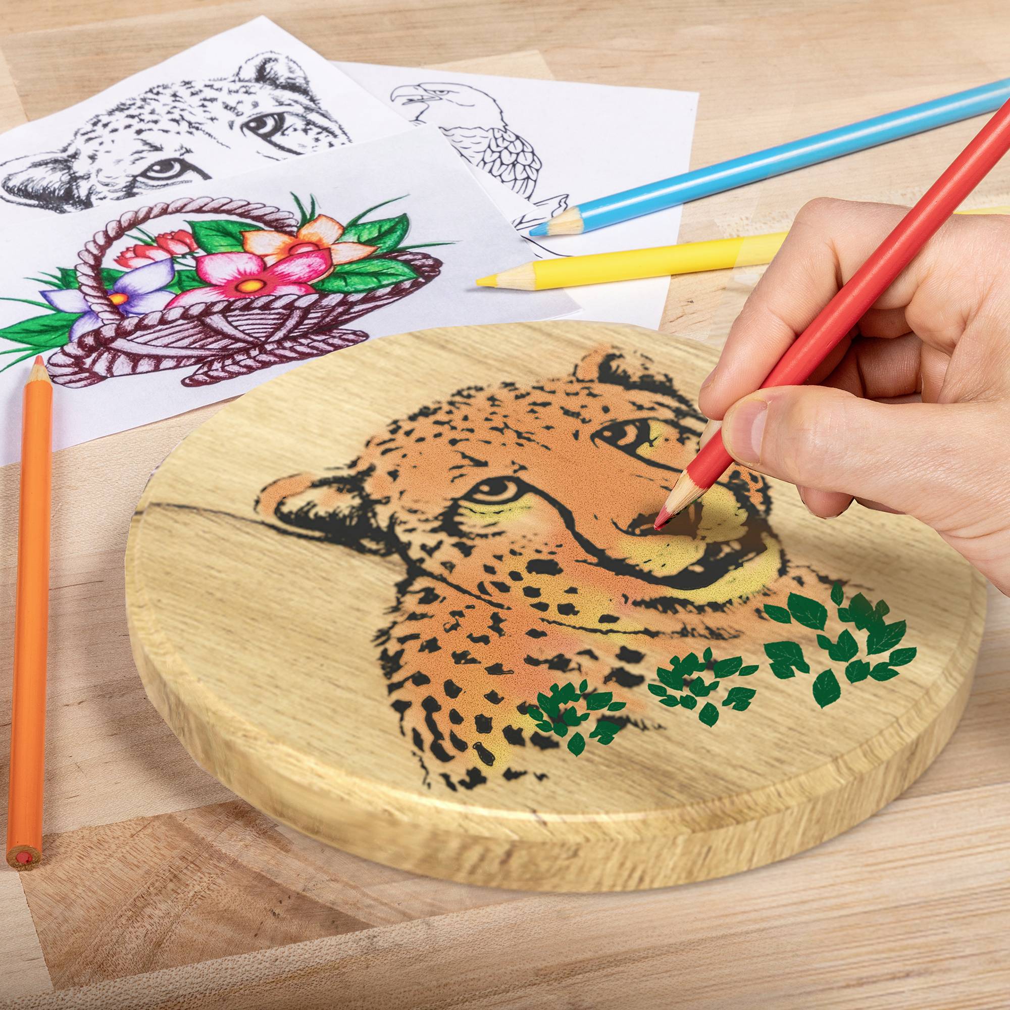 Person zeichnet mit rotem Stift ein Gepard-Gesicht auf Holzbrett, umgeben von Buntstiften und Tierzeichnungen auf Papier.