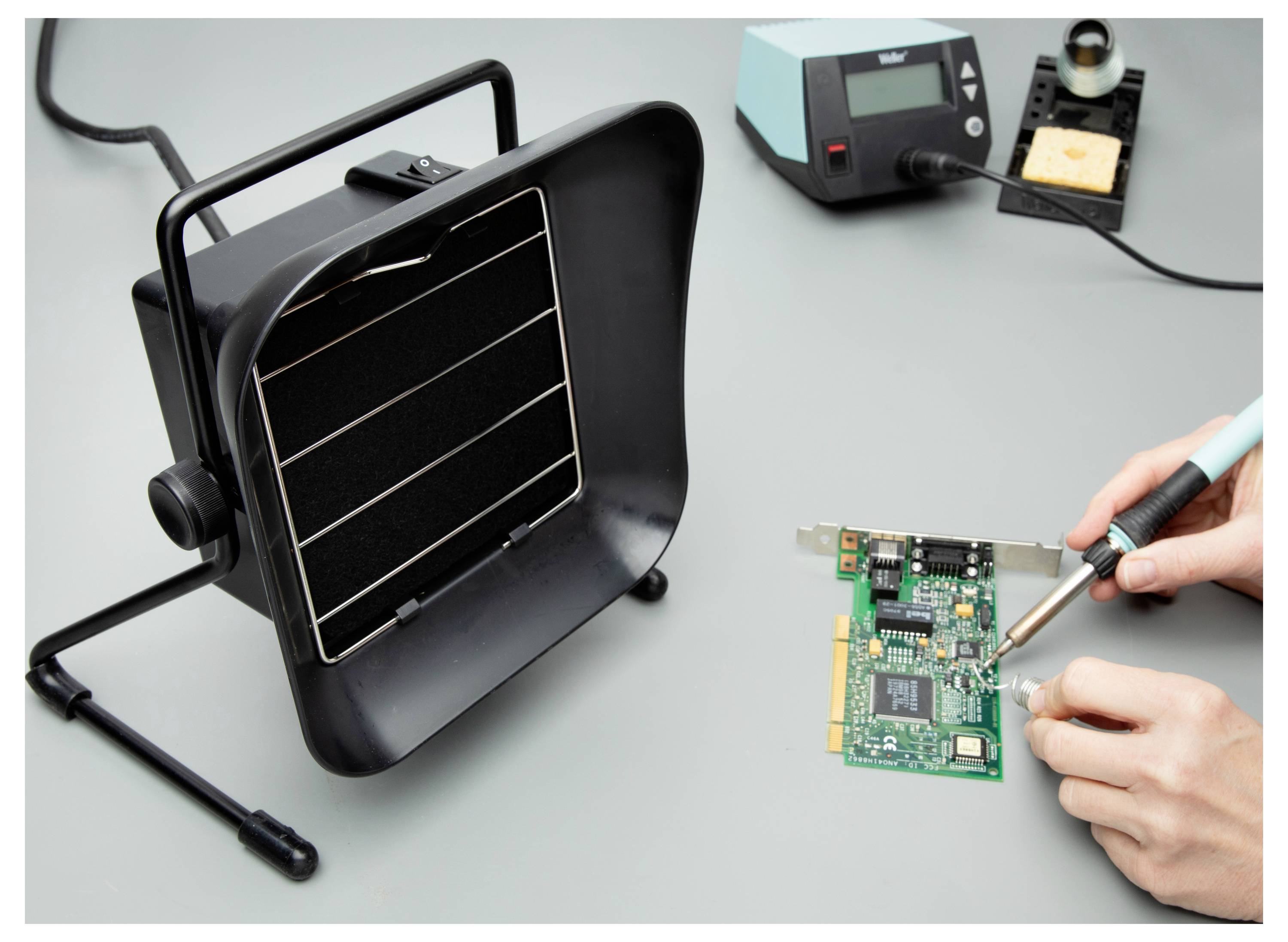 Eine Person lötet eine Leiterplatte mit einem Lötkolben, wobei sie einen Absauganlage für die Sicherheit verwendet. Ein elektronisches Reparaturaufbau wird gezeigt.