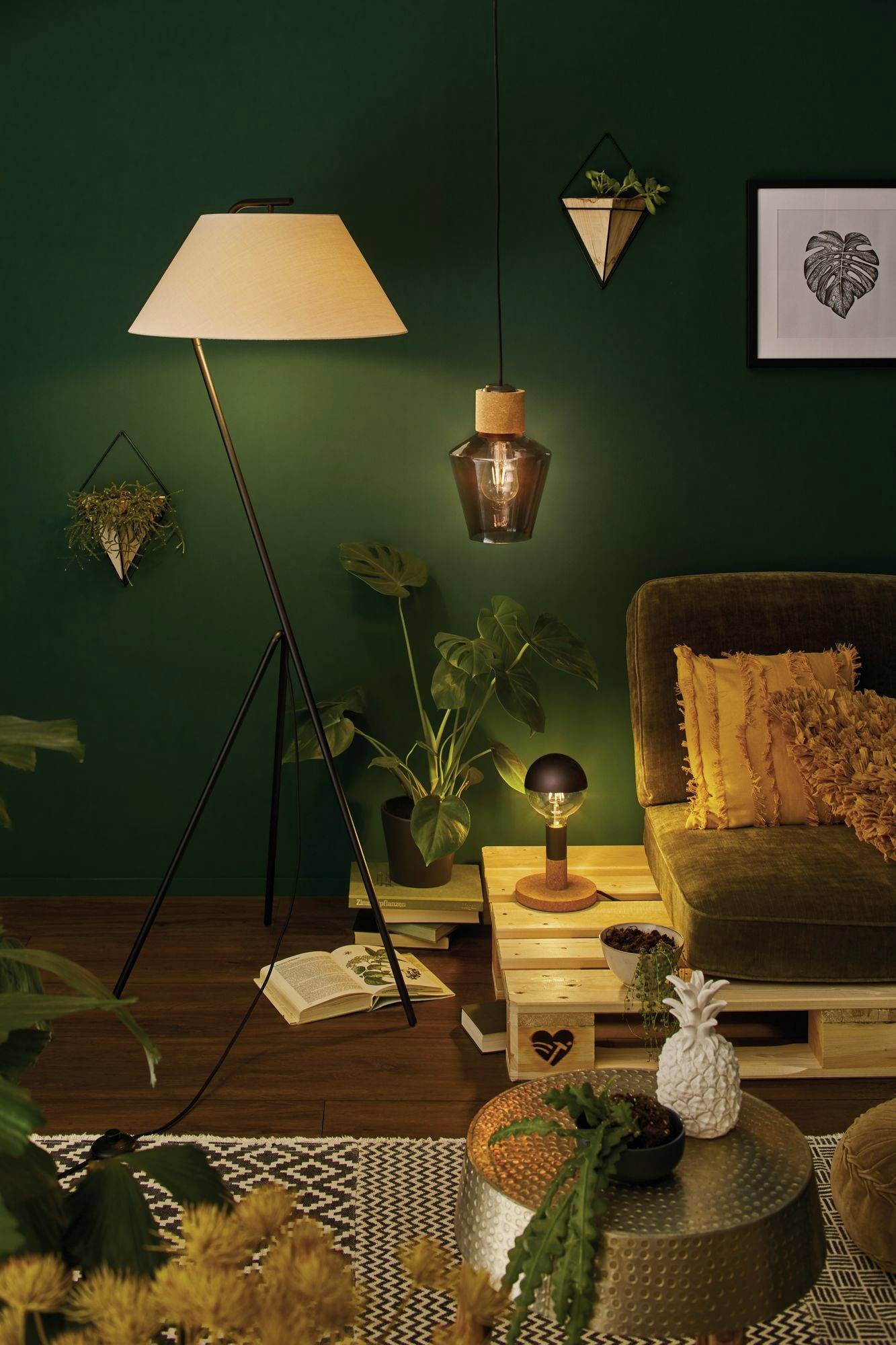 Gemütliches Wohnzimmer mit grünem Sofa, Pflanzen und warmem Licht von einer Stehlampe und Hängeleuchte, Bücher auf dem Boden.