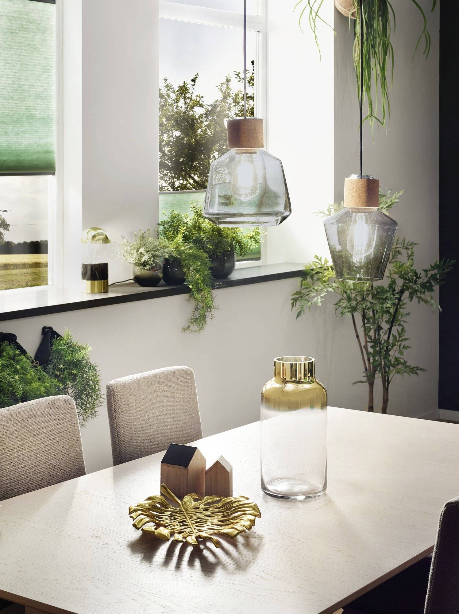 Esszimmer mit hellem Holztisch, dekoriert mit Vase, Holzfiguren und Blatt-Schale. Hängelampen und Pflanzen am Fenster im Hintergrund.