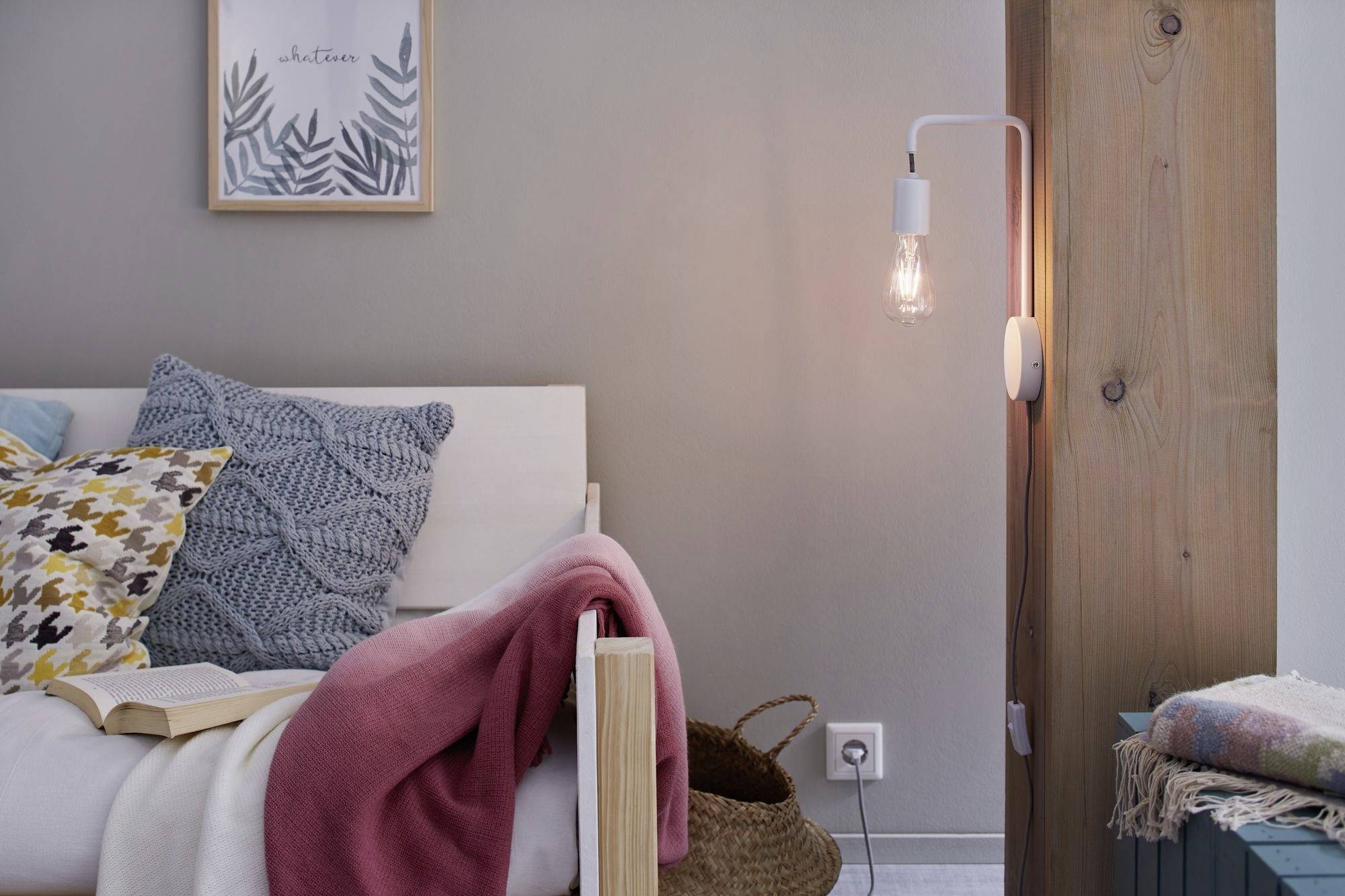 Gemütliches Schlafzimmer mit einem Bett, grauen und bunten Kissen, einer rosa Decke, einem Buch und einer Wandlampe neben Holzbalken.