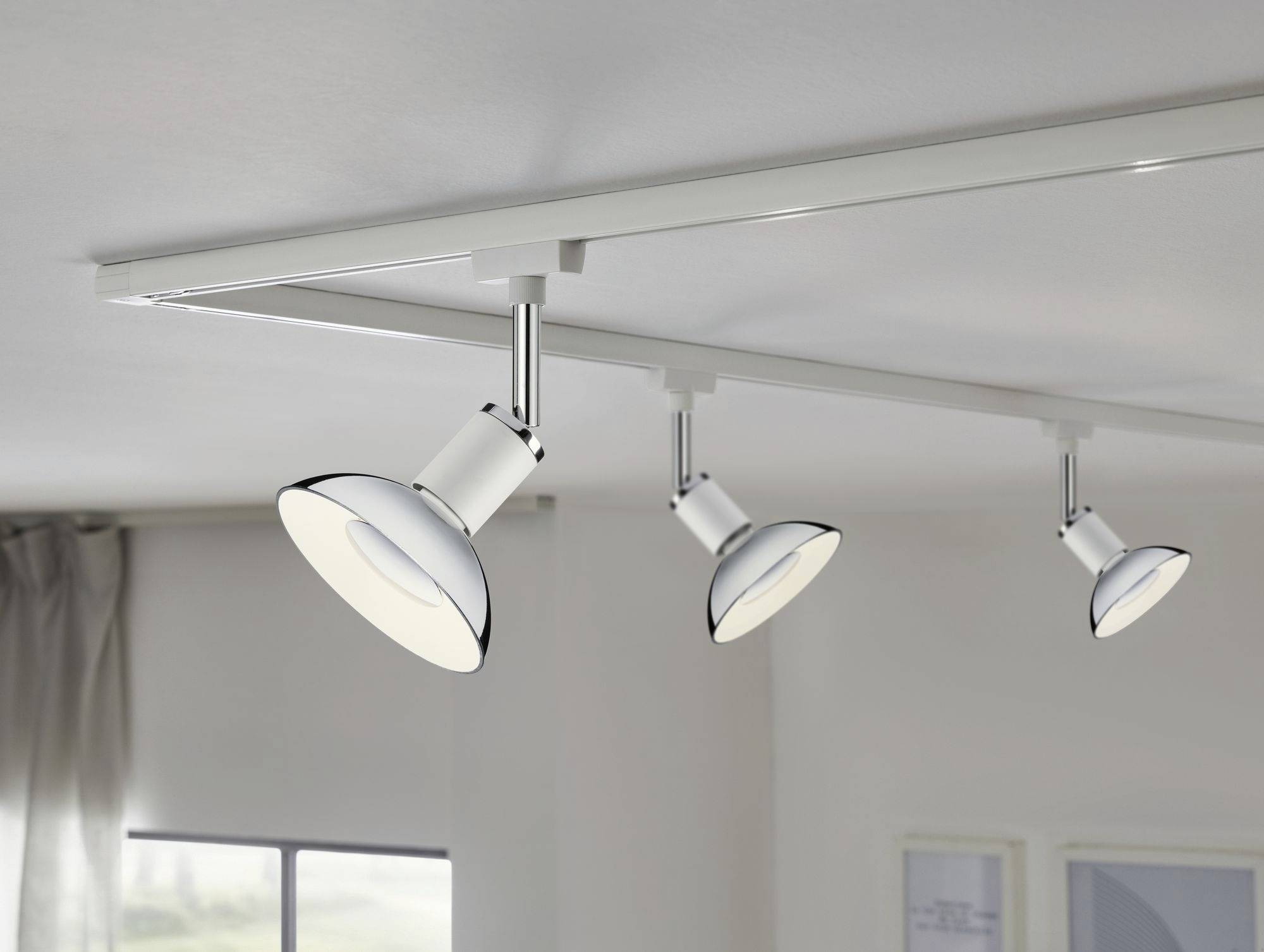 Drei Deckenstrahler an einer Schiene leuchten einen Raum aus. Ein Fenster mit Vorhang ist im Hintergrund sichtbar.