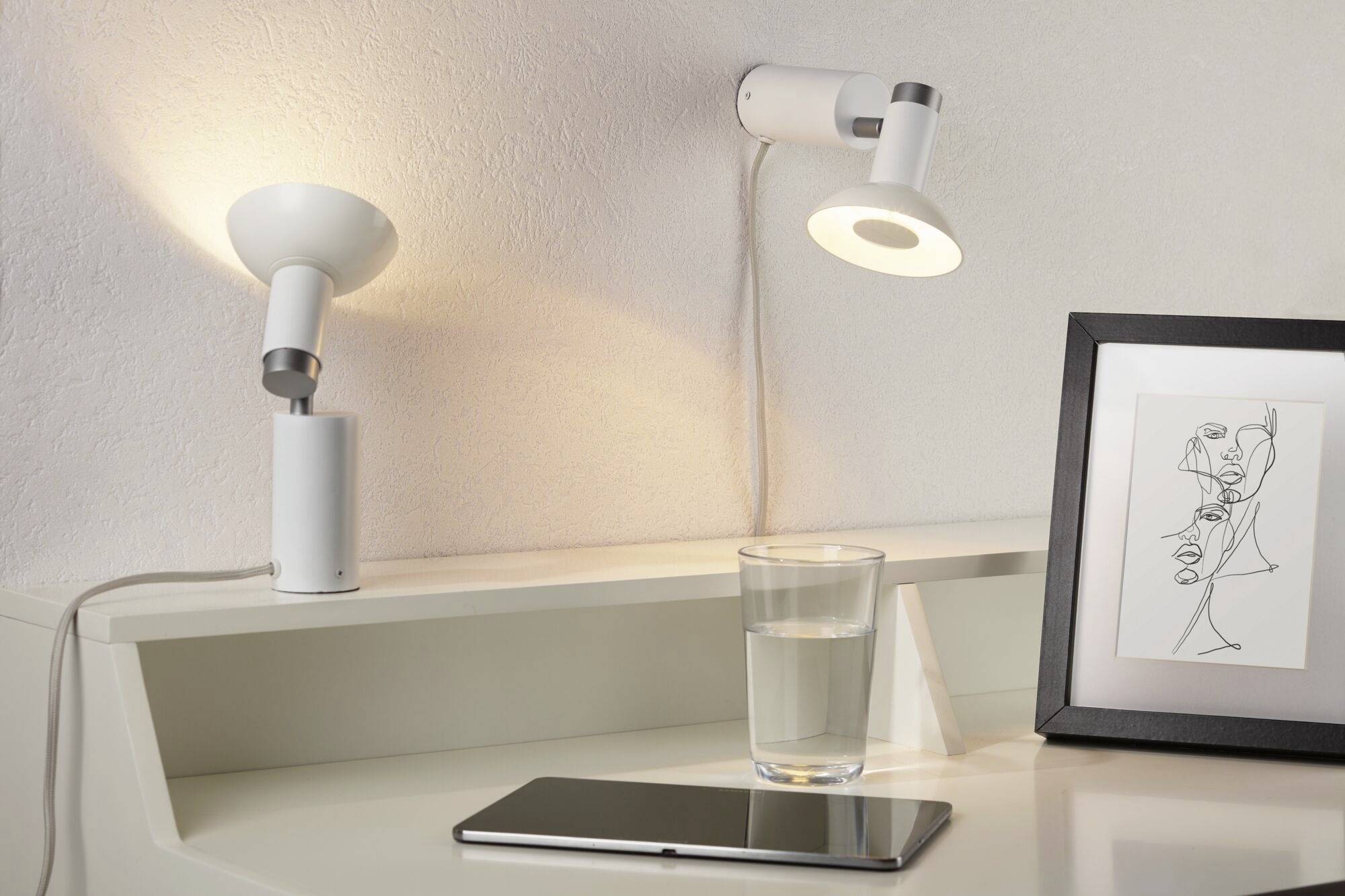 Schreibtisch mit zwei Lampen, ein Glas Wasser, ein Smartphone und ein gerahmtes Kunstwerk. Stimmung einer modernen, minimalistischen Arbeitsfläche.