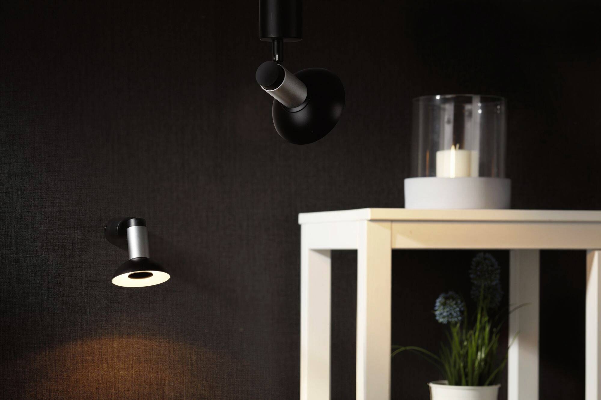 Eine schwarze Wandlampe leuchtet einen Teil der Wand an. Rechts steht ein weißer Tisch mit einer Pflanze und einer Kerze im Glas.