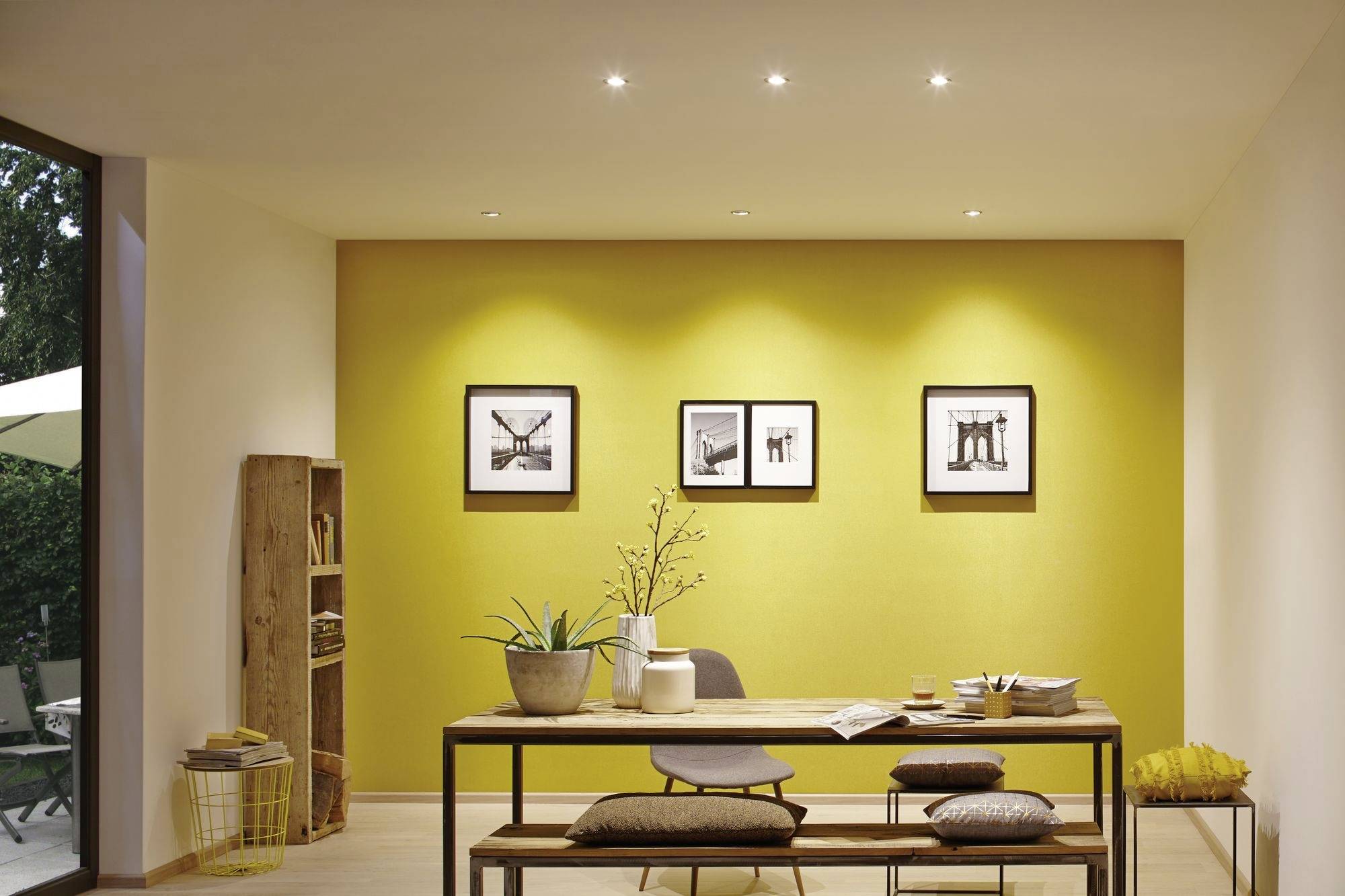 Ein modern eingerichtetes Esszimmer mit gelber Akzentwand, Holztisch, Bänken, Regalen und mehreren Bildern an der Wand.