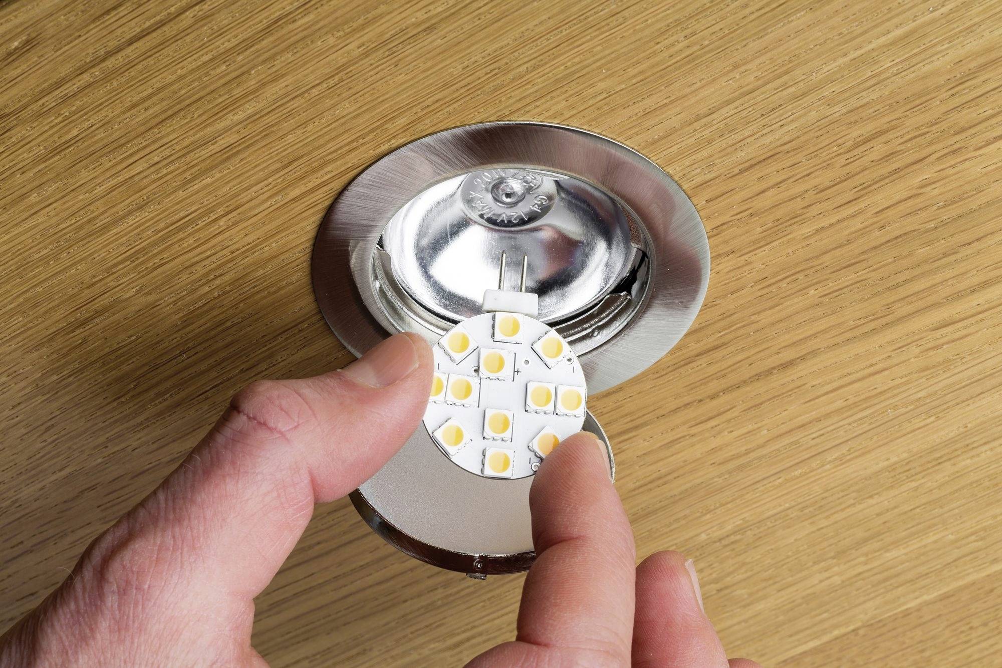 Person hält eine runde LED-Lampe über einer offenen Deckenleuchte, bereit zum Einsetzen auf einer hölzernen Oberfläche.