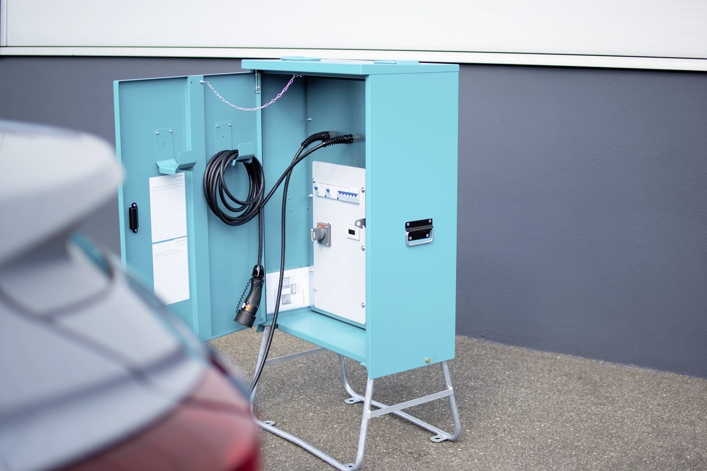 Ein offener, türkisfarbener Kasten mit Kabeln und Elektronik, vermutlich eine Ladestation, steht auf einem grauen Boden neben einem Fahrzeug.