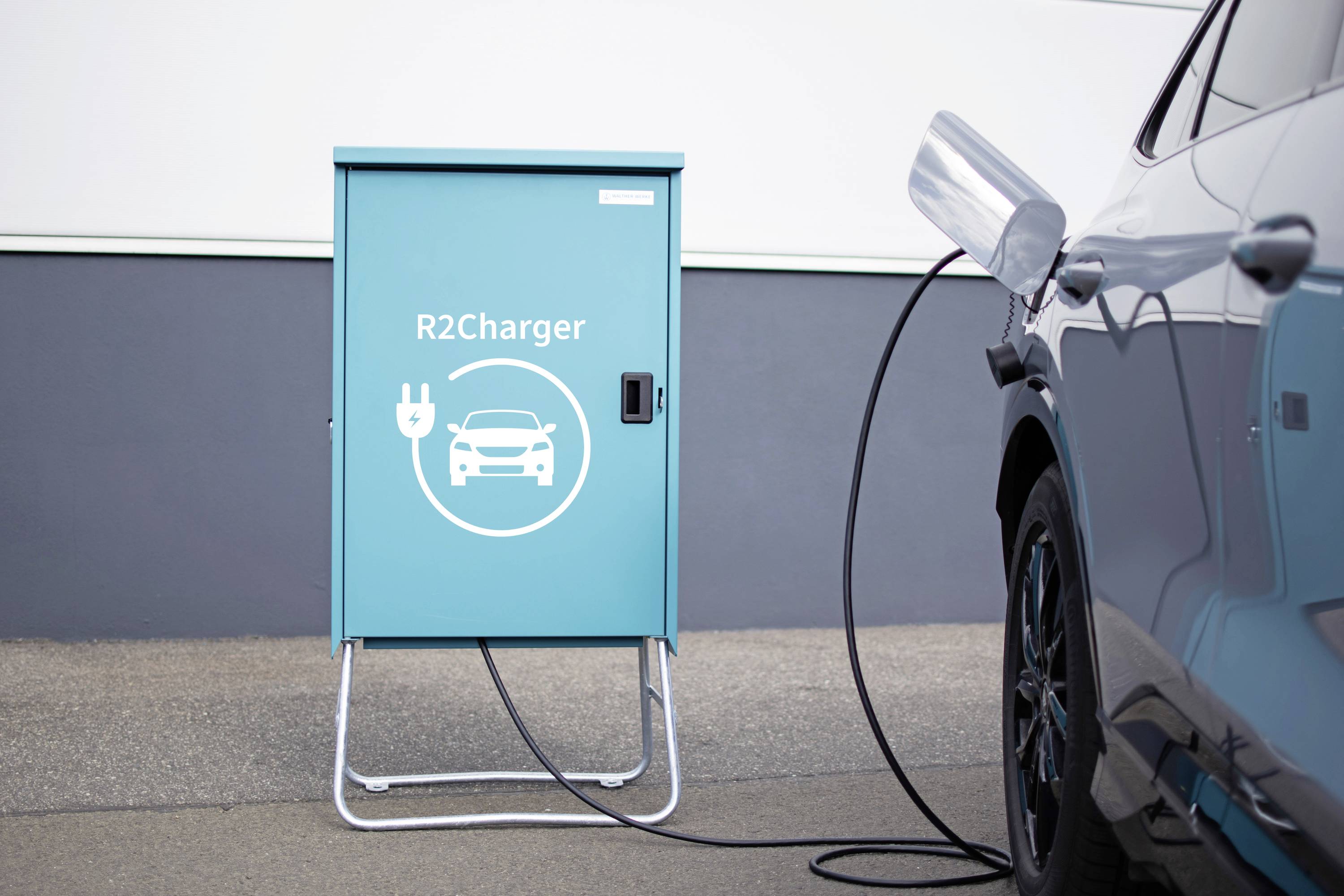 Ein Elektroauto wird über ein Ladekabel mit einer Ladestation namens 'R2Charger' verbunden. Der Standort ist städtisch.