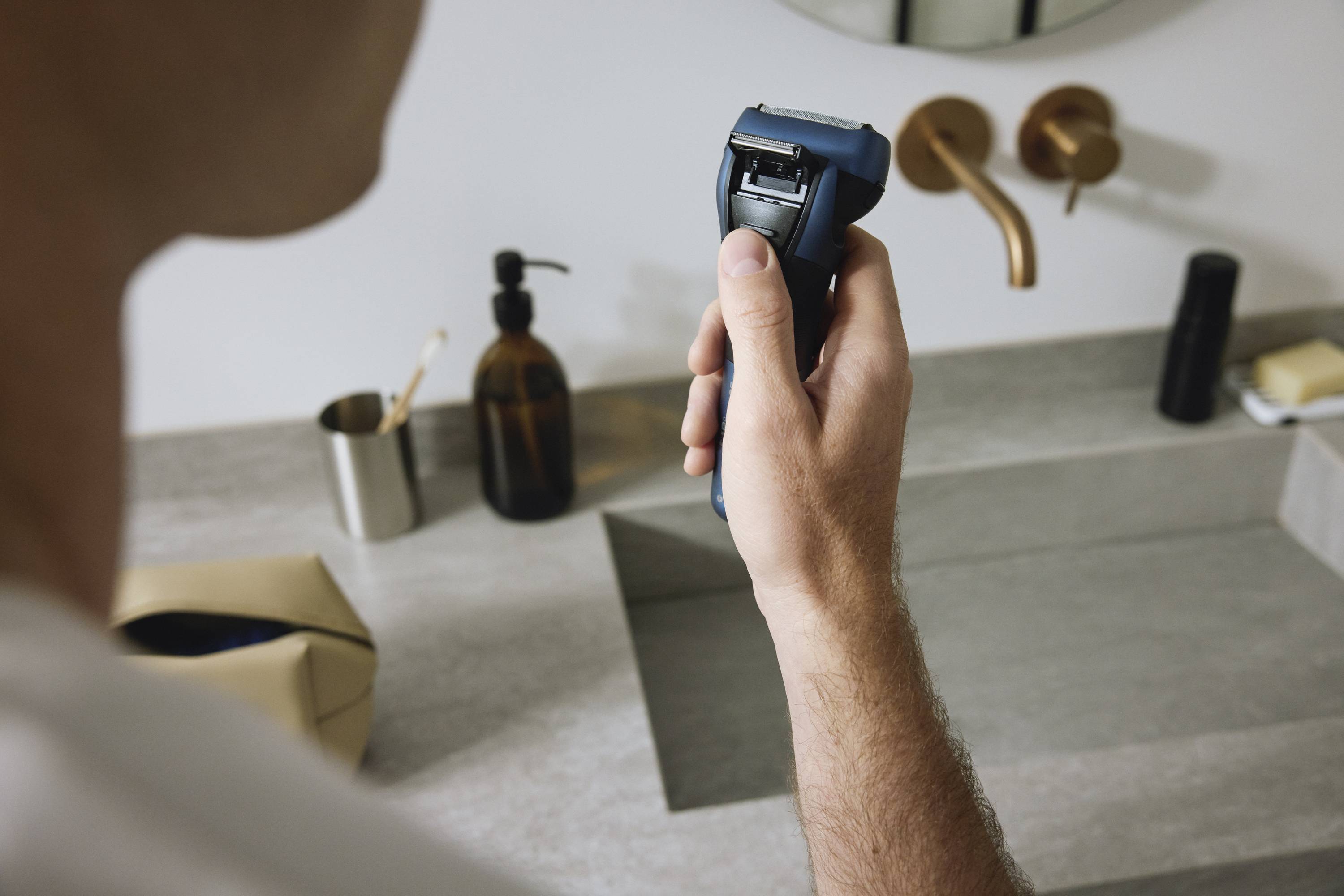 Person hält einen elektrischen Rasierer über einem Waschbecken in einem modernen Badezimmer. Im Hintergrund Seifenspender und Wasserhähne.