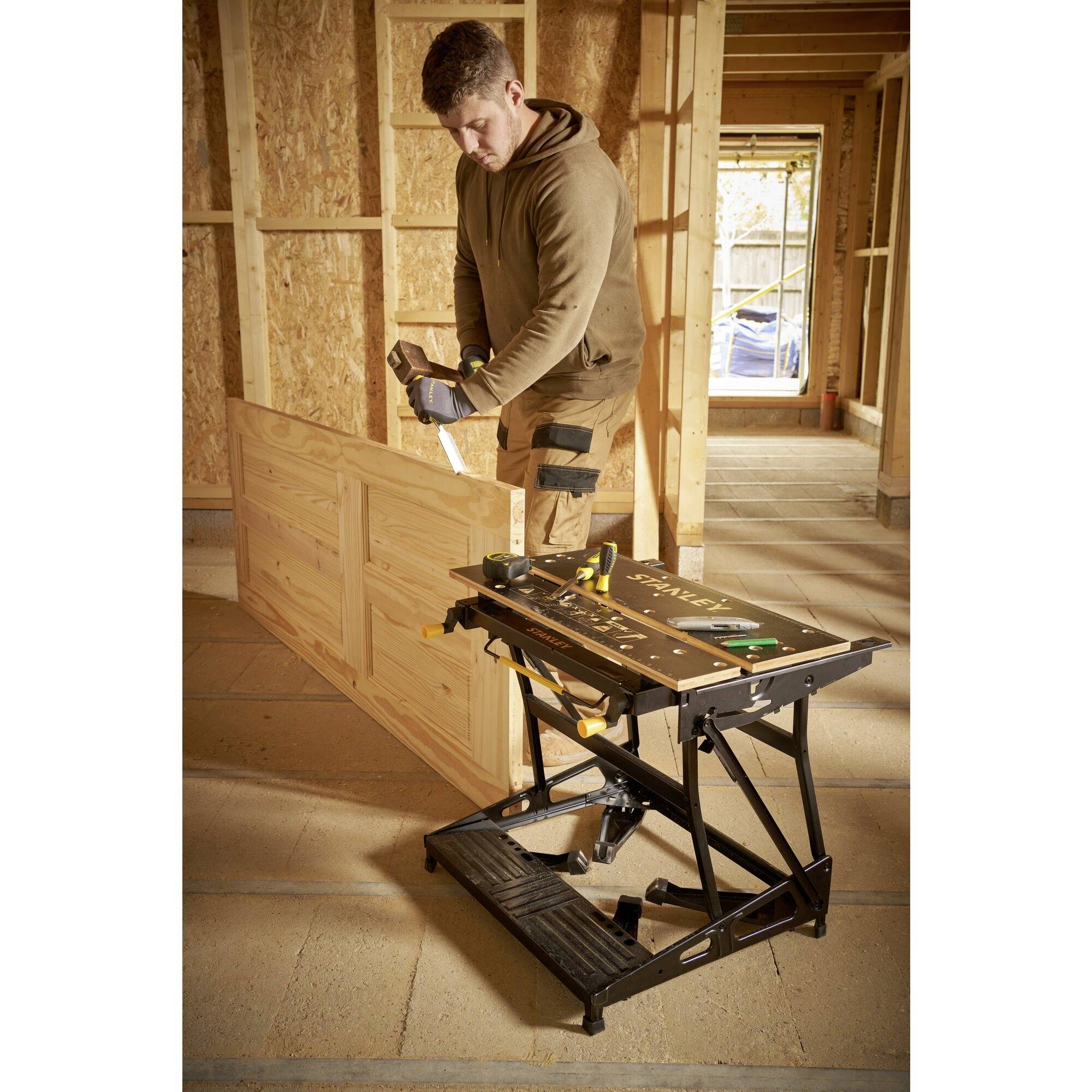 Ein Mann in Arbeitskleidung sägt in einer Werkstatt ein Holzbrett mit einer Handsäge. Eine Werkbank mit Werkzeugen steht rechts.