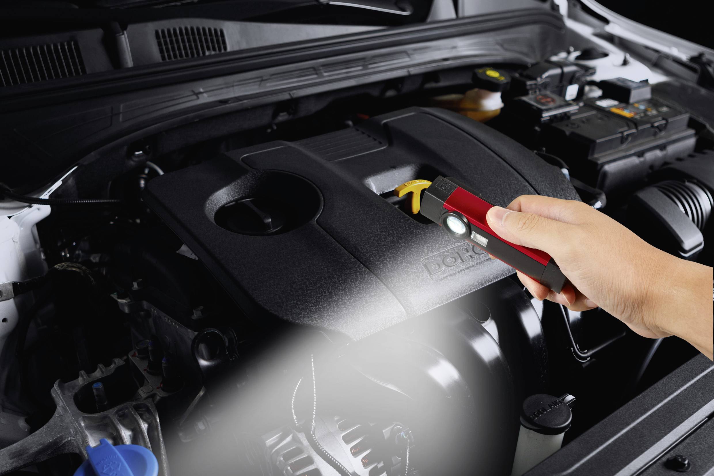 Person überprüft den Motor eines Autos mit einer Taschenlampe. Die Motorhaube ist geöffnet, und der Fokus liegt auf dem Motorblock.