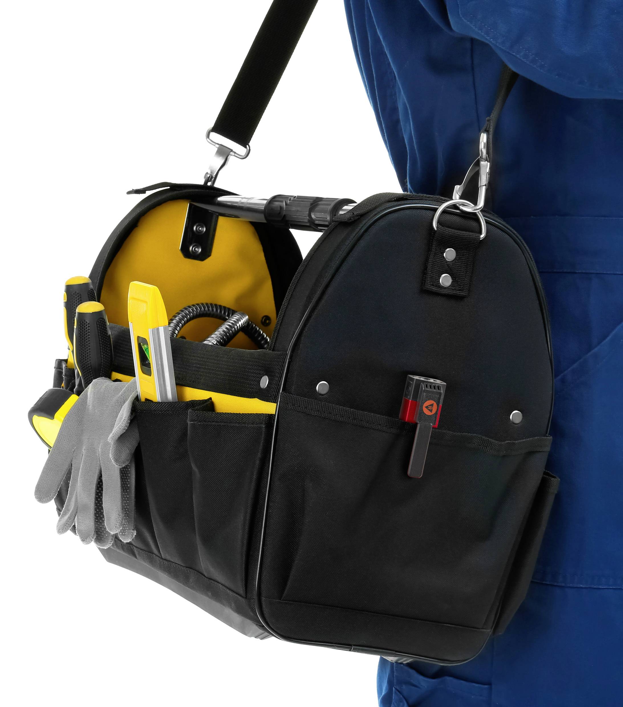 Handwerker in blauer Arbeitskleidung trägt eine schwarze Werkzeugtasche voller Handwerkzeuge, wie Schraubendreher und Zangen.