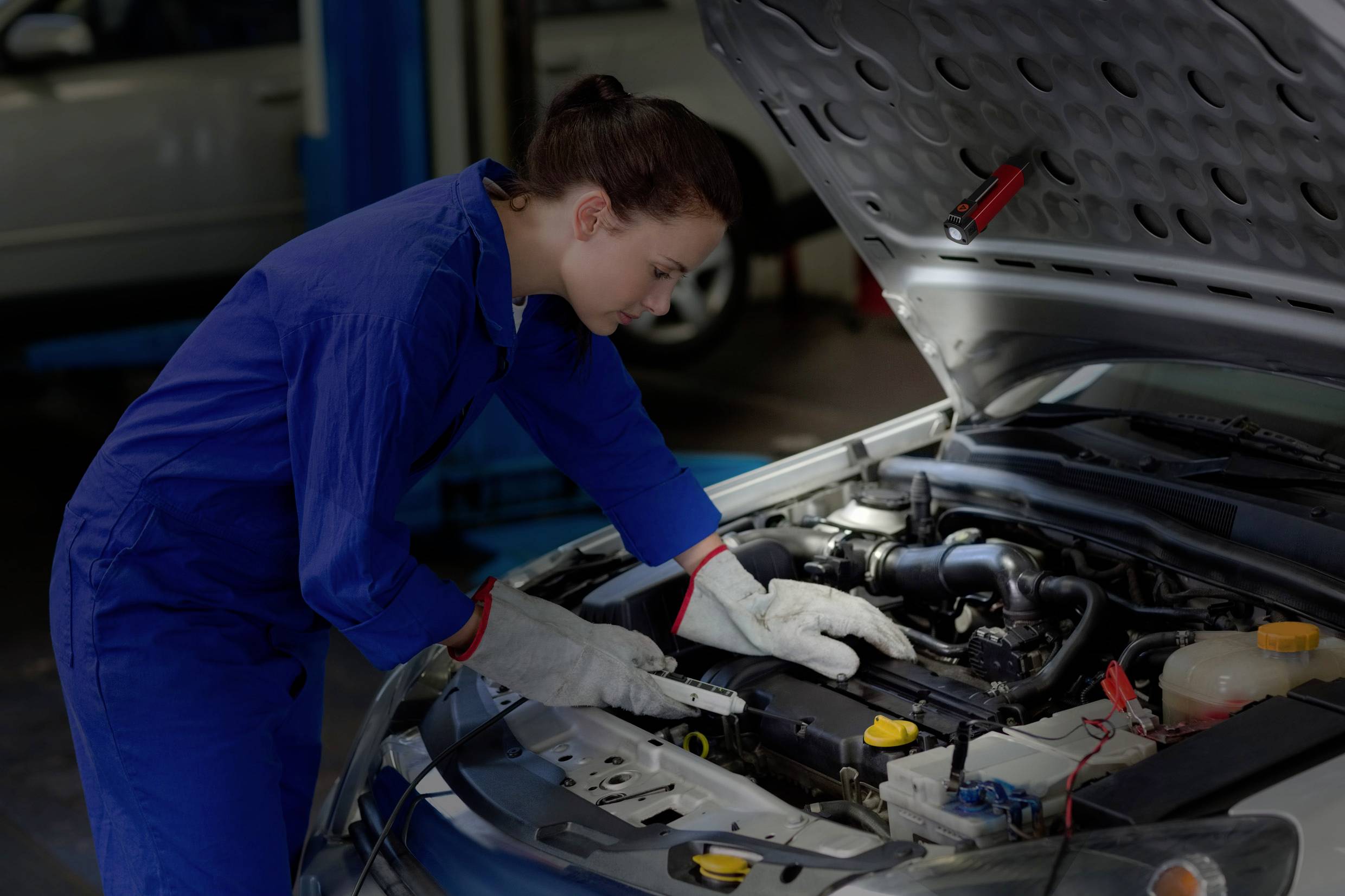 Eine Mechanikerin in blauer Arbeitskleidung repariert den Motor eines Autos in einer Werkstatt, fokussiert und mit Werkzeugen arbeitend.