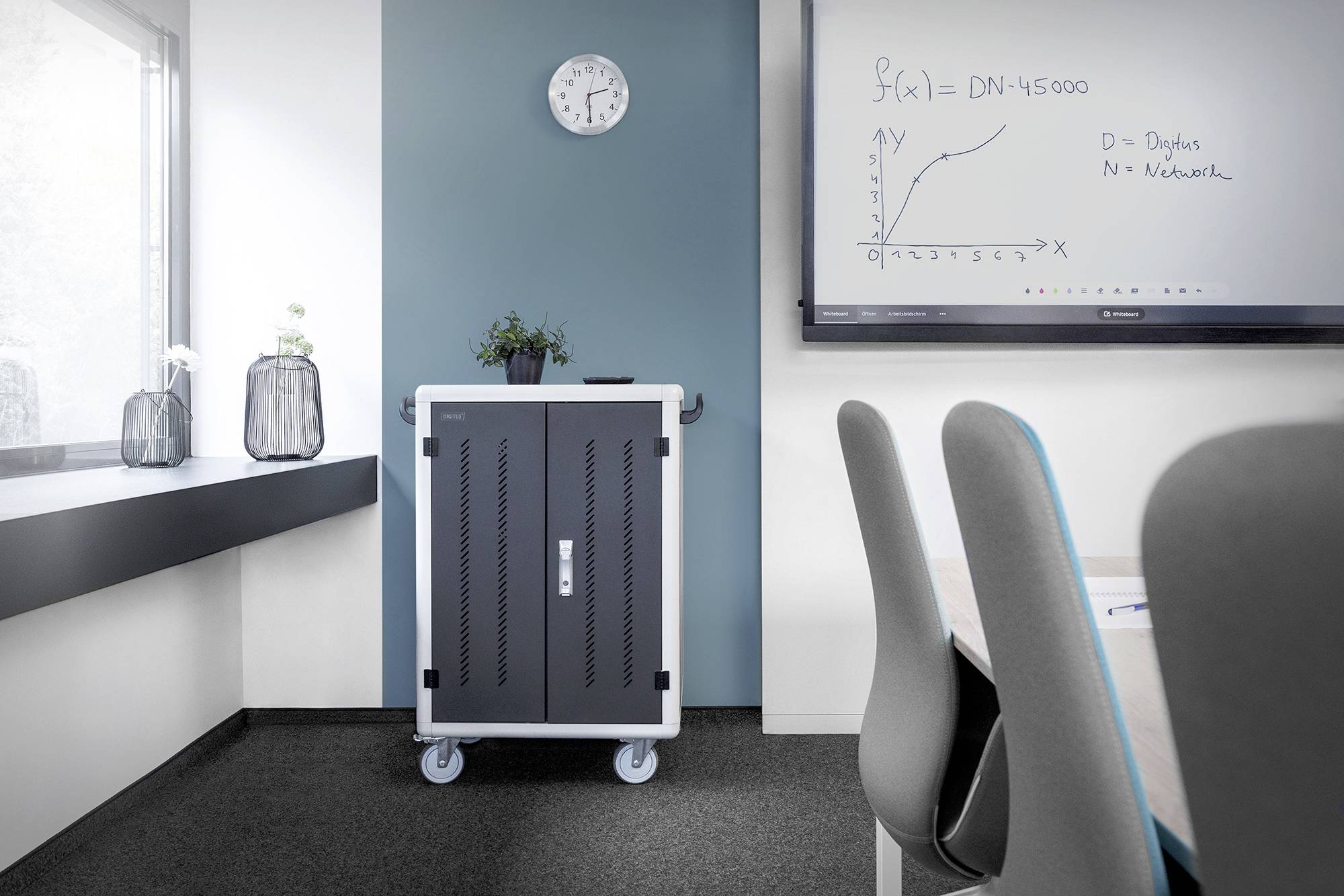 Ein Konferenzraum mit einem Tisch und Stühlen, einem Rollschrank, einer Wandtafel mit einer gezeichneten Funktion und einer Uhr an der Wand.