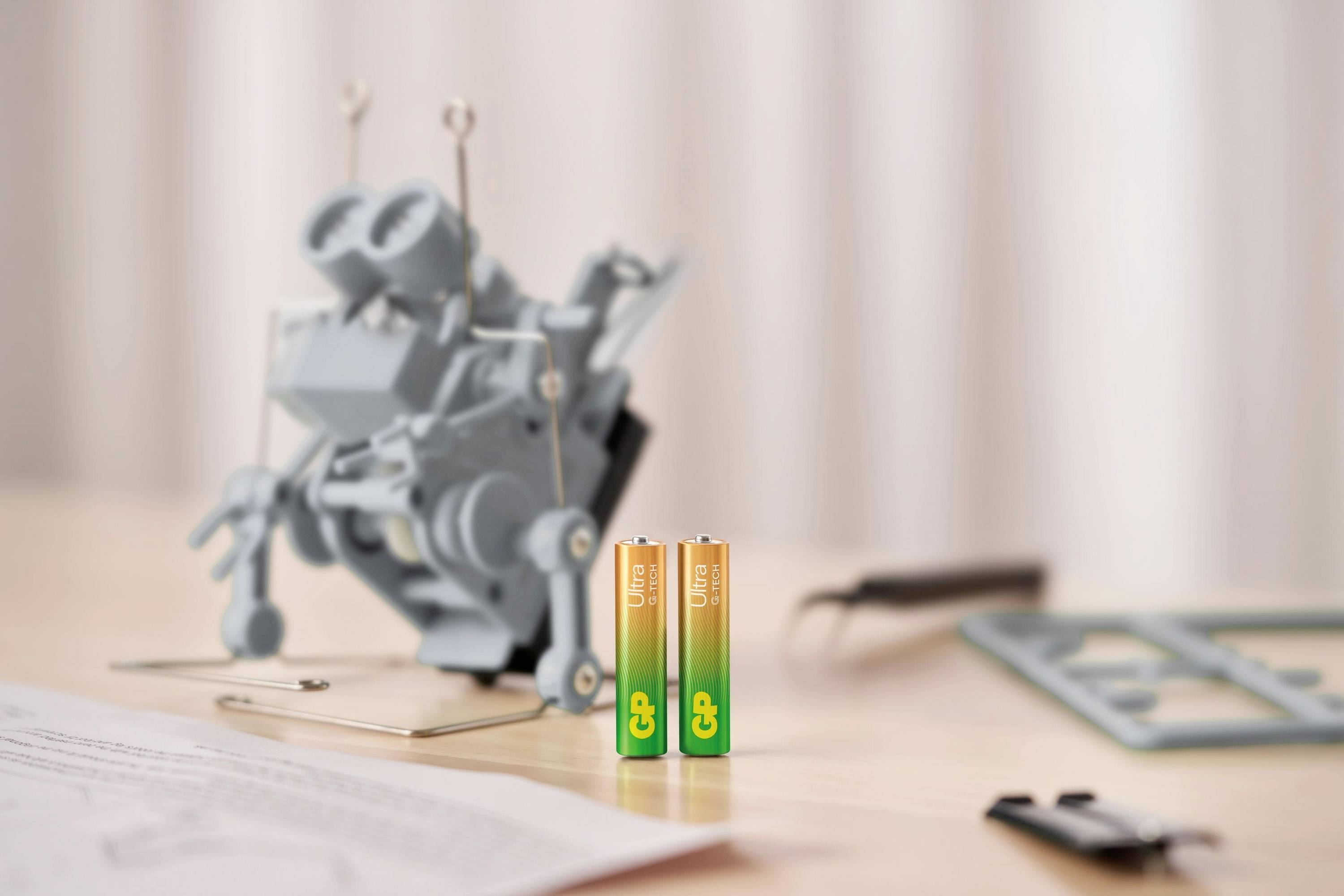 Zwei AA-Batterien im Vordergrund; unscharf im Hintergrund liegt eine zerlegte Kamera oder ein mechanisches Gerät auf einem Tisch.