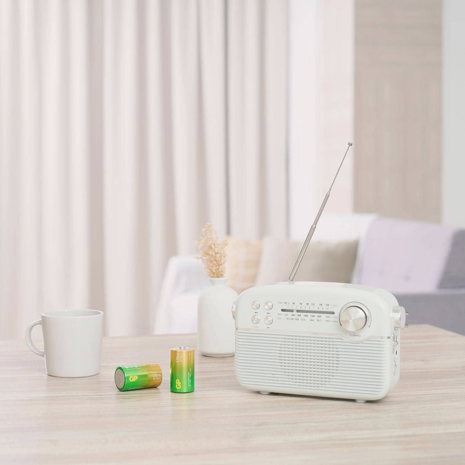 Weißes Retro-Radio auf einem Tisch neben Tasse und zwei Batterien in einem hellen Wohnraum.