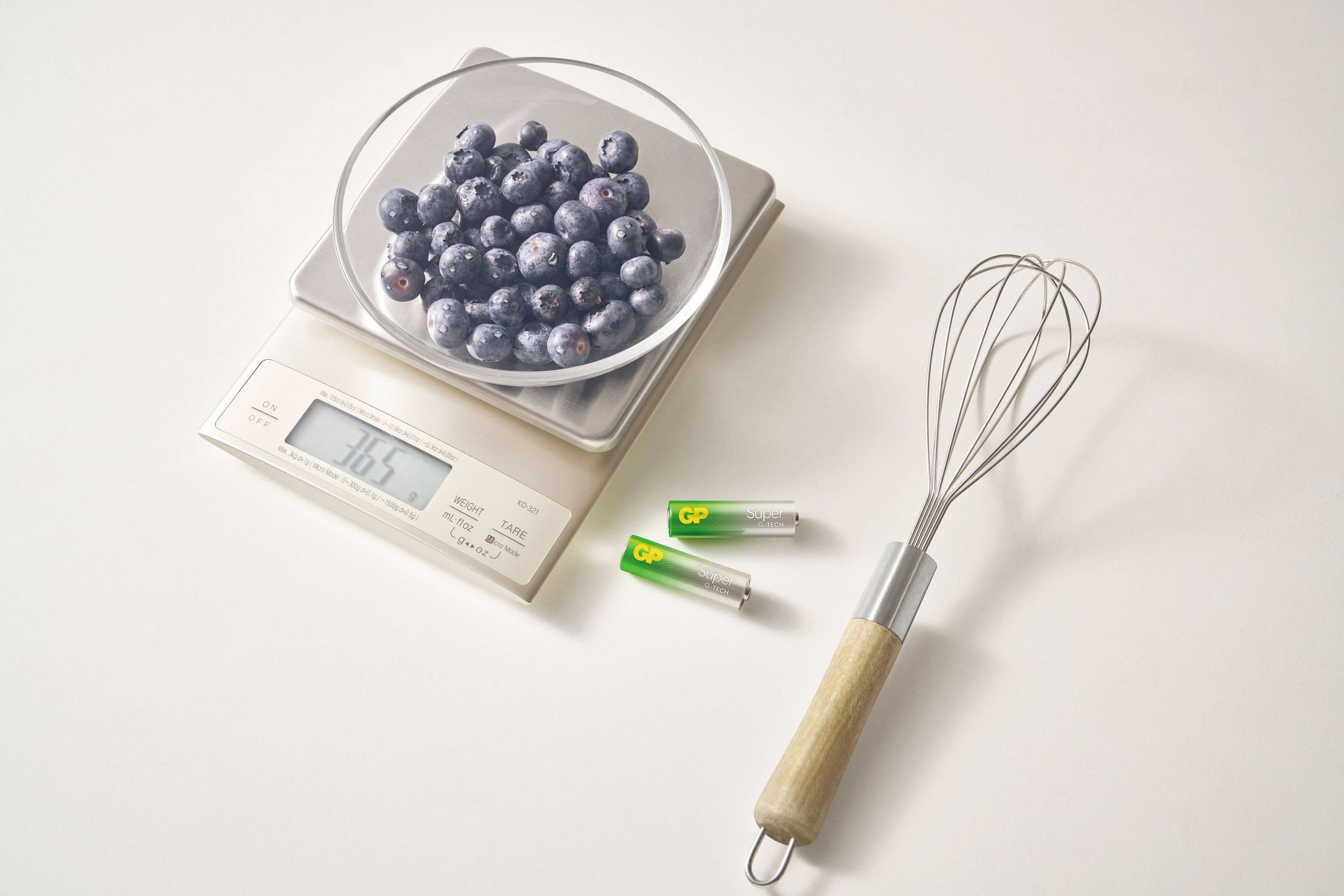 Schüssel mit Blaubeeren auf einer Küchenwaage, daneben Schneebesen und zwei Batterien auf hellem Untergrund.