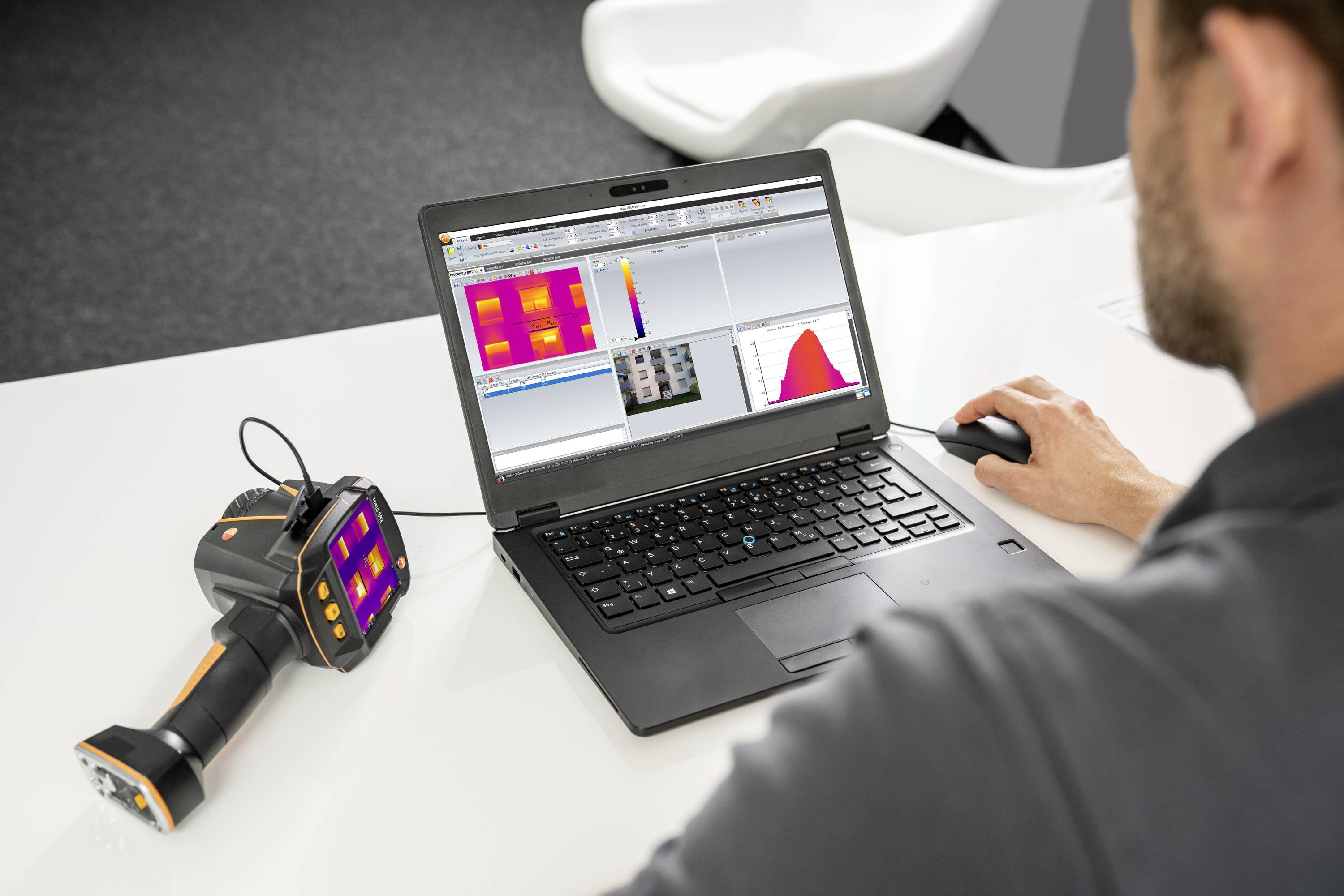 Ein Mann analysiert Wärmebilder auf einem Laptop. Ein Handgerät, vermutlich ein Thermografiekamera, liegt daneben auf dem Tisch.