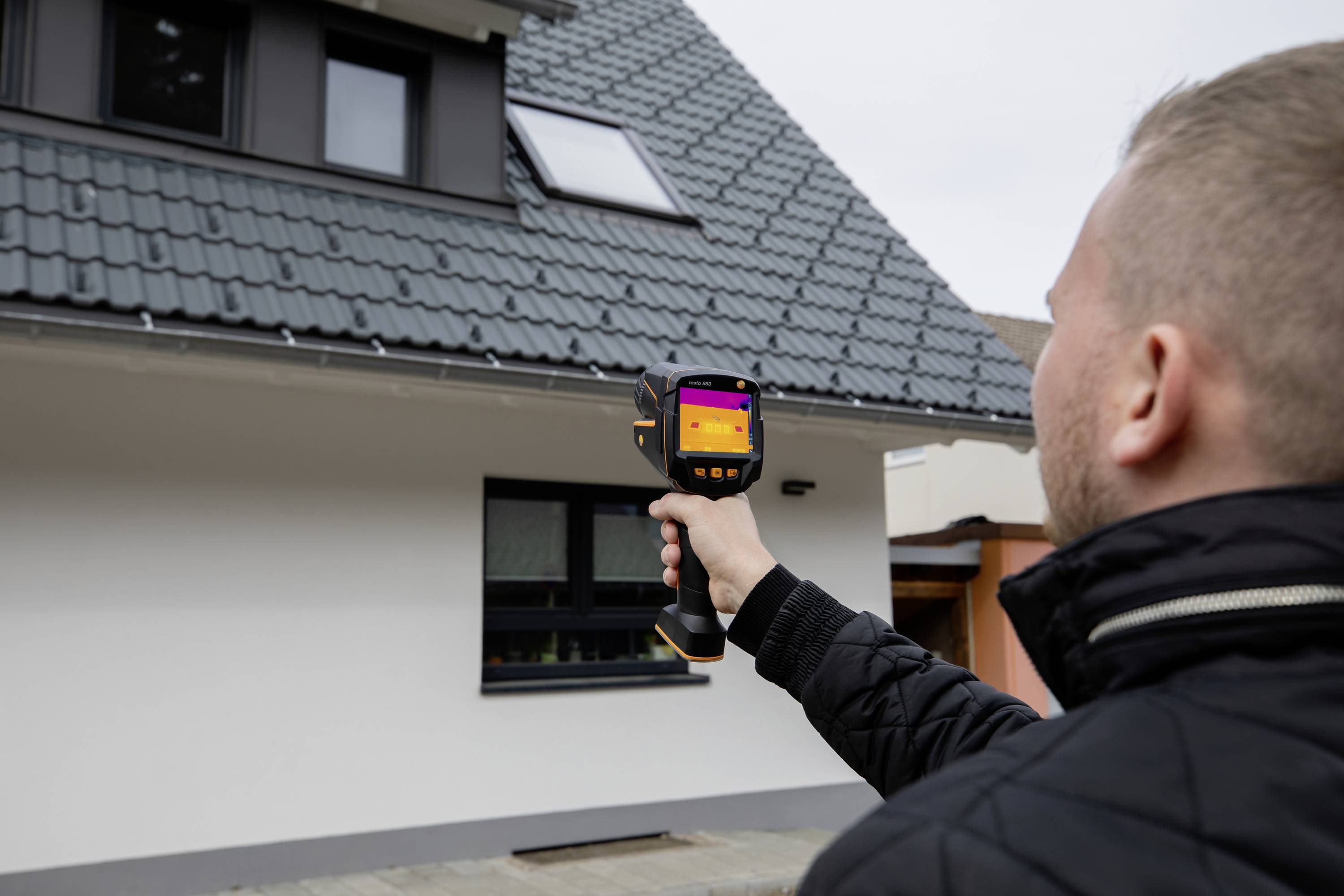 Ein Mann verwendet ein Wärmebildkamera-Gerät, um ein Haus zu inspizieren. Die Kamera zeigt Temperaturunterschiede auf der Fassade.