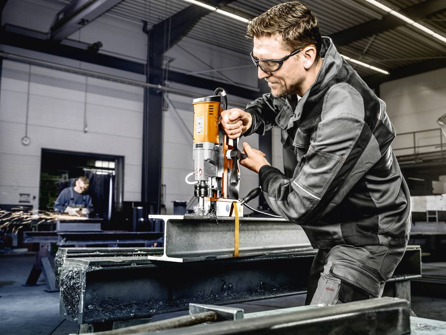 Ein Mann in Arbeitskleidung bedient eine magnetische Bohrmaschine auf einem Stahlträger in einer Werkstatt. Im Hintergrund arbeitet eine weitere Person.