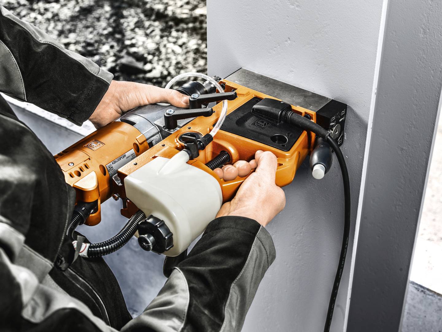 Person nutzt eine orangefarbene Bohrmaschine, um in ein graues Metallträger zu bohren. Im Hintergrund ist unscharf eine Baustelle zu sehen.