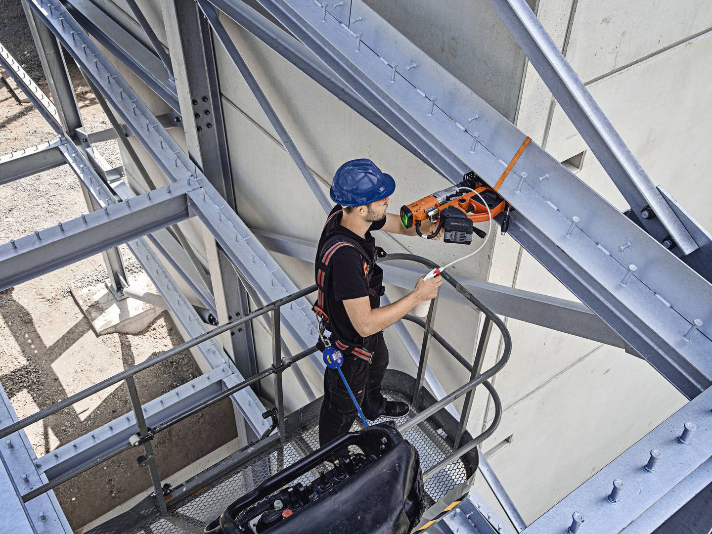Ein Bauarbeiter im blauen Helm arbeitet mit einem elektrischen Werkzeug auf einem Gerüst, befestigt mit Sicherheitsgurten an Stahlträgern.