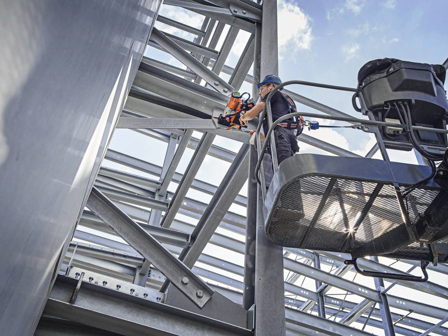 Ein Arbeiter in Schutzkleidung steht auf einer Hebebühne und verwendet ein Elektrowerkzeug, um an einer großen Stahlkonstruktion zu arbeiten.