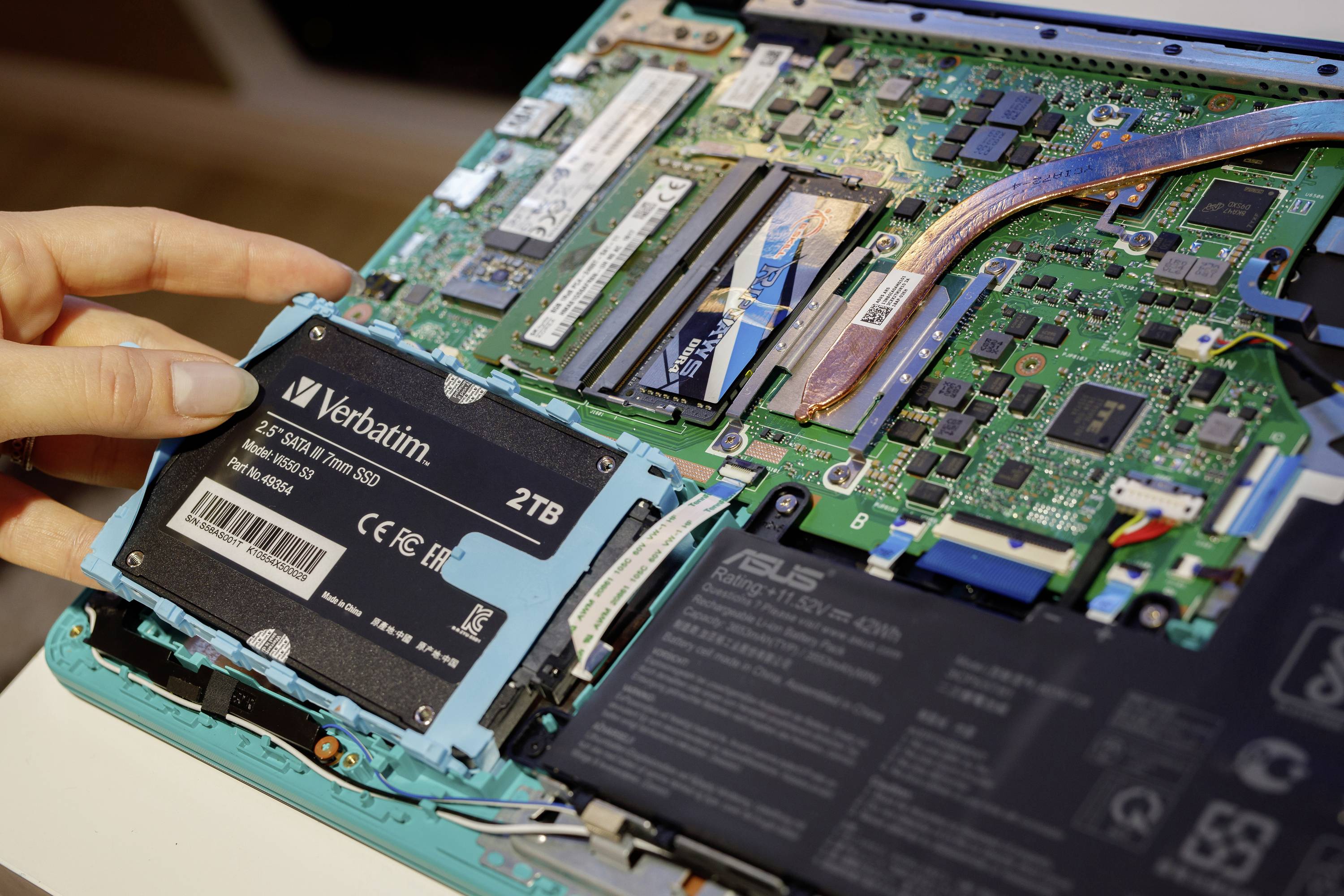 Eine Hand installiert eine SSD-Festplatte in einen Laptop, dessen Gehäuse offen ist und die interne Elektronik sichtbar macht.