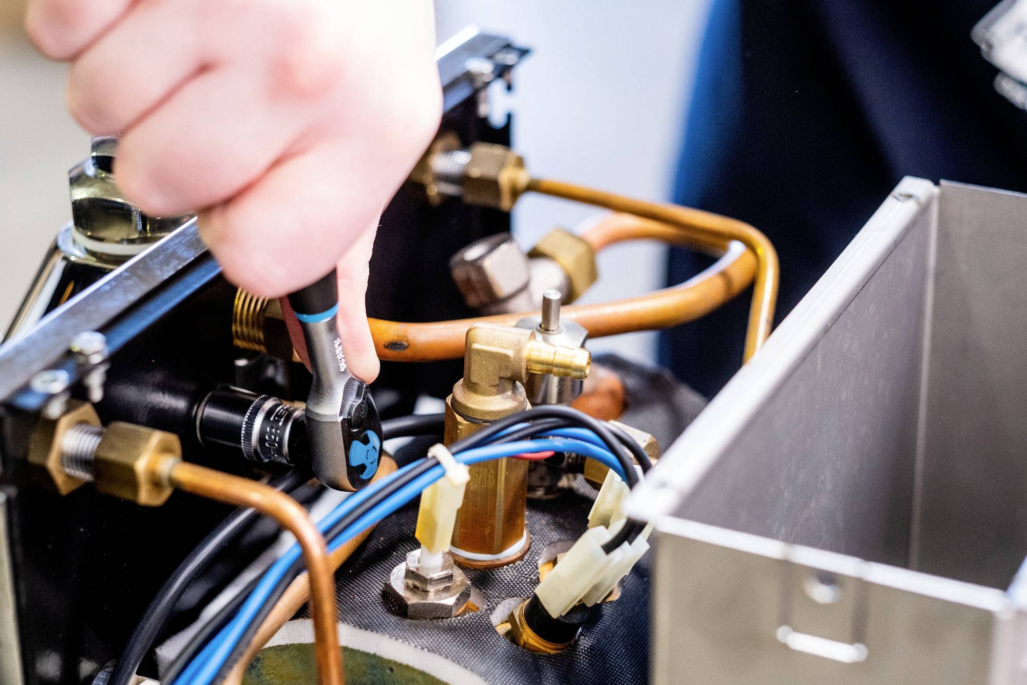 Eine Person verwendet einen Ratschenschlüssel, um eine Metallkomponente zwischen Kupferrohren und elektrischen Leitungen in einem mechanischen Gerät zu befestigen.