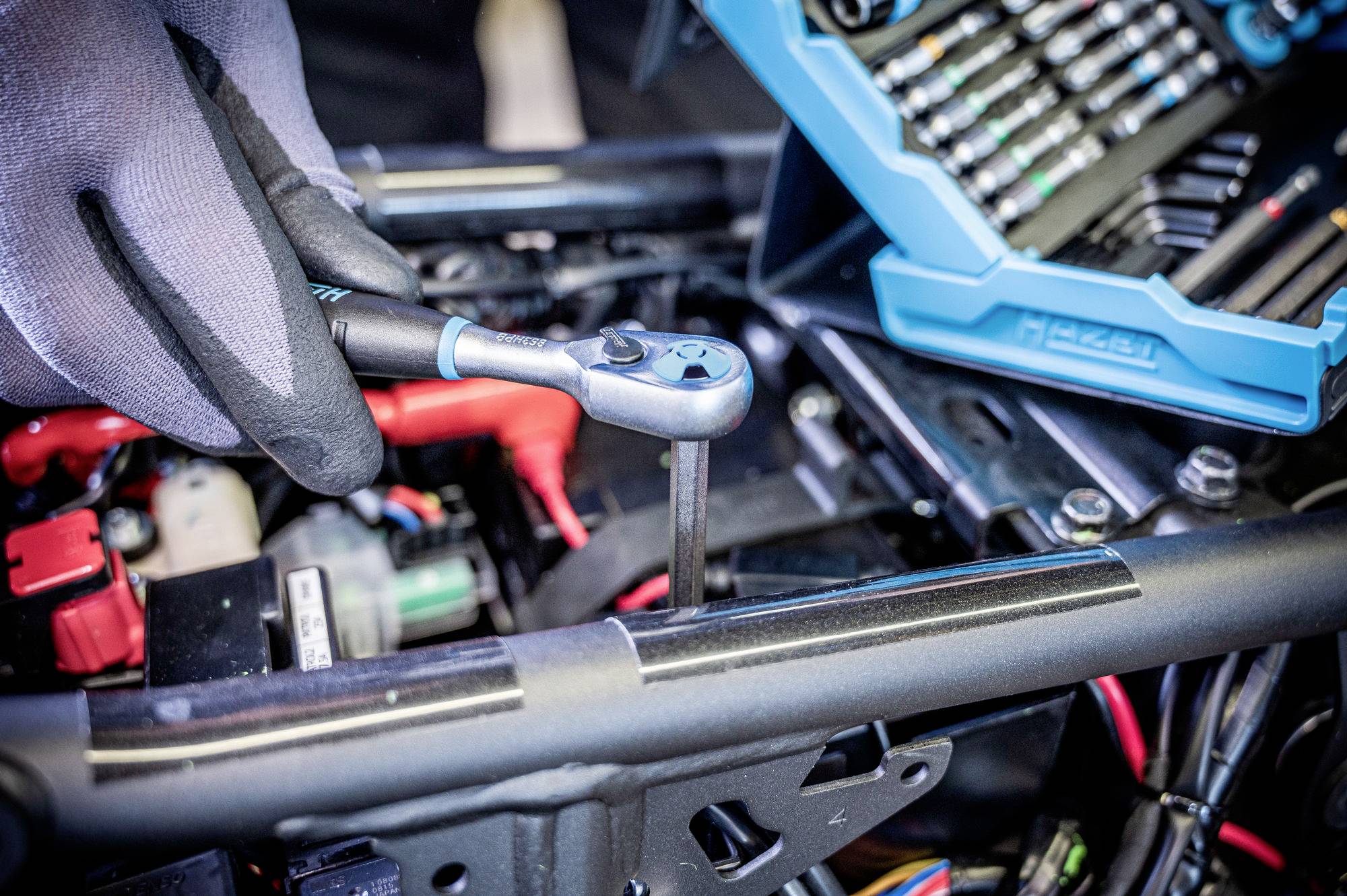 Eine Person mit Handschuhen benutzt einen Ratschenschlüssel, um einen Bolzen an einer mechanischen Anlage zu festigen, während im Hintergrund eine Werkzeugkiste zu sehen ist.