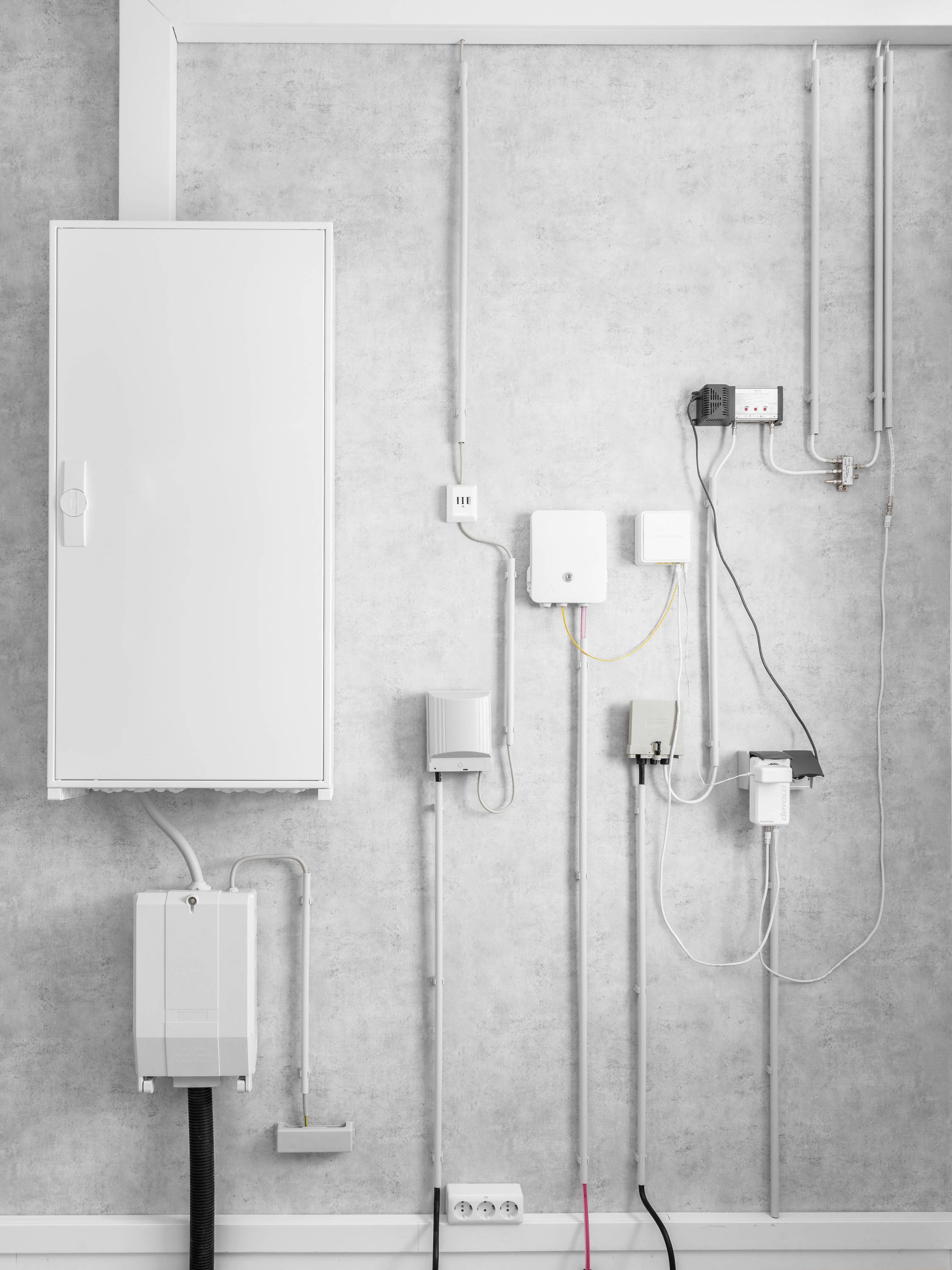 Elektroinstallationen an einer grauen Wand, einschließlich eines großen weißen Schaltschrankes und diverser Verteilerkästen mit angeschlossenen Kabeln.