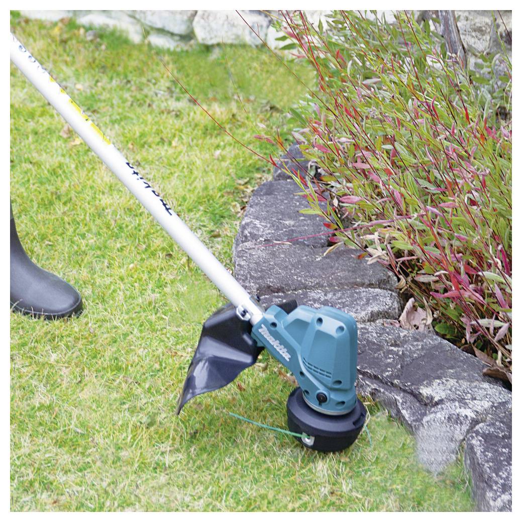 Eine Person benutzt einen batteriebetriebenen Fadentrimmer zum Rasenmähen neben einem Gartenbeet mit Pflanzen und trägt schwarze Stiefel zum Schutz.