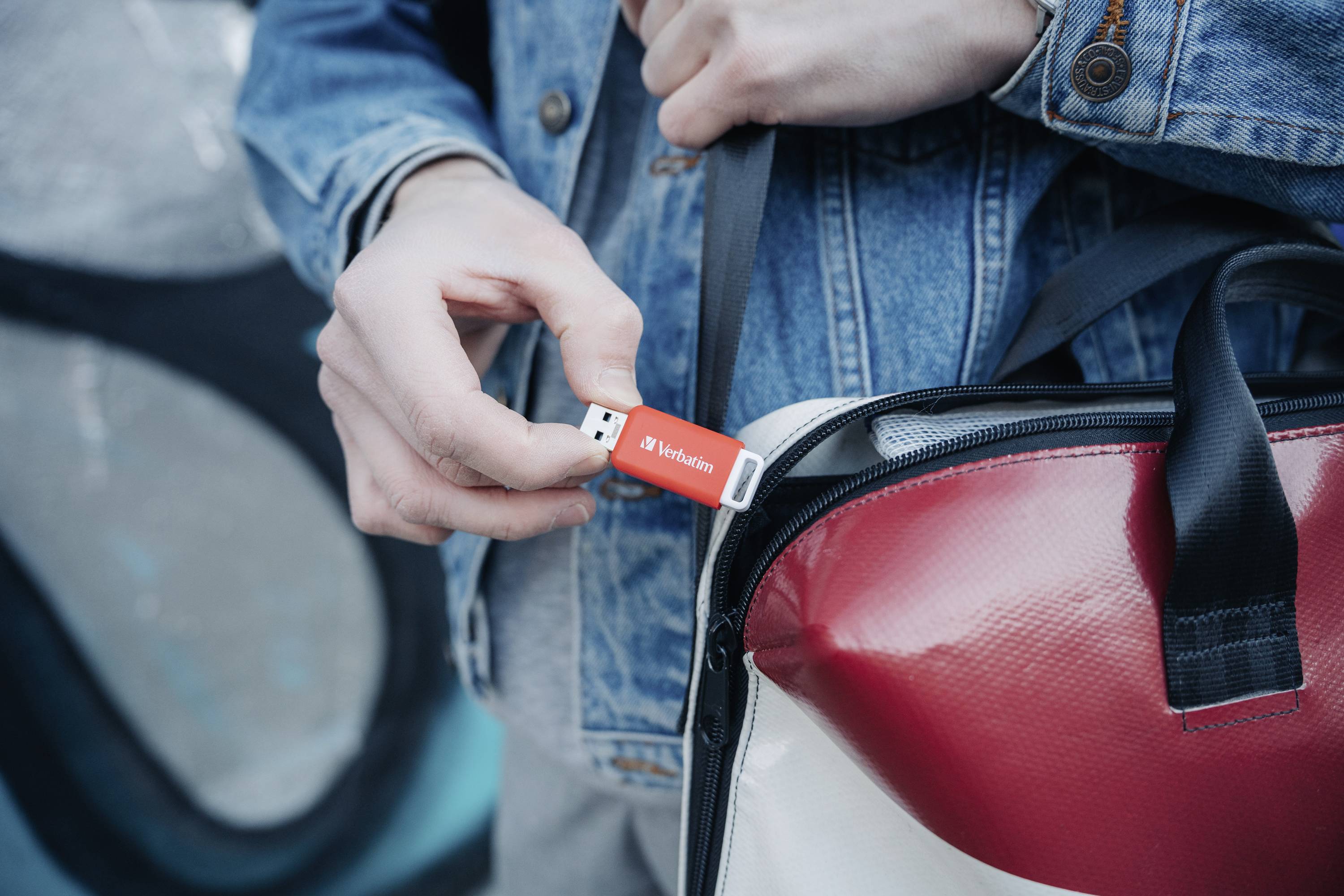 Eine Person steckt einen roten USB-Stick in eine Tasche. Sie trägt eine Jeansjacke. Der USB-Stick trägt die Aufschrift 'Verbatim'.