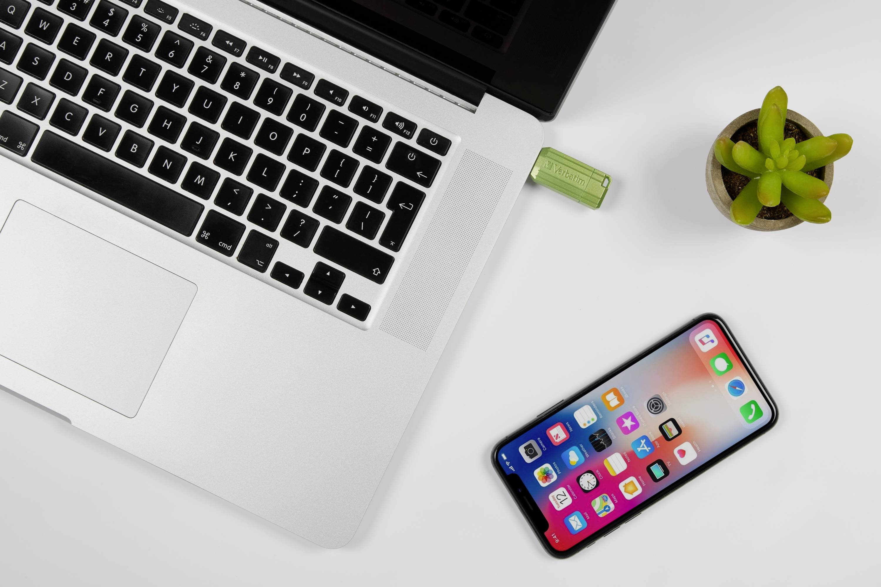 Laptop mit USB-Stick oben links, Smartphone mit Apps rechts, kleine grüne Pflanze oben rechts. Arbeitsbereich auf weißem Tisch.